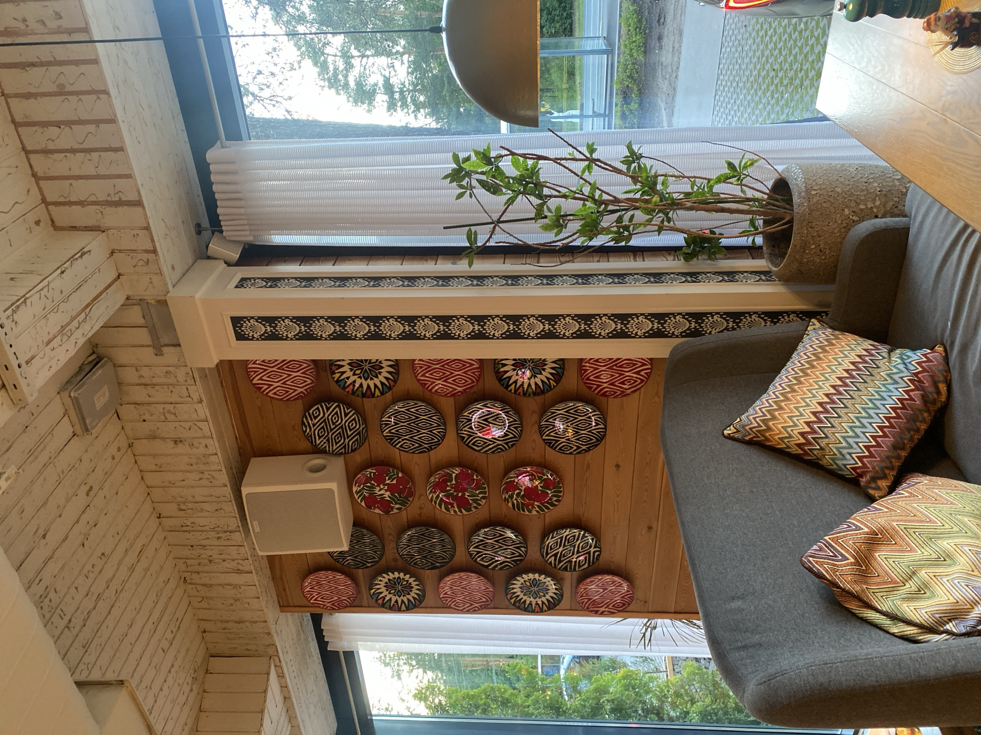 The image depicts the interior of a room with a wooden paneled wall adorned with numerous decorative plates, a gray sofa with two colorful zigzag-patterned pillows, and a tall potted plant. A large window with white curtains reveals an outdoor scene with trees and a bus stop. The ceiling is made of white-painted wooden planks, and a white speaker is mounted on the wall. In the foreground, a wooden table with a salt and pepper grinder and a small figurine is visible. The overall ambiance suggests a cozy, possibly bohemian, interior design. The location is identified as Solnechnoye, Russia.