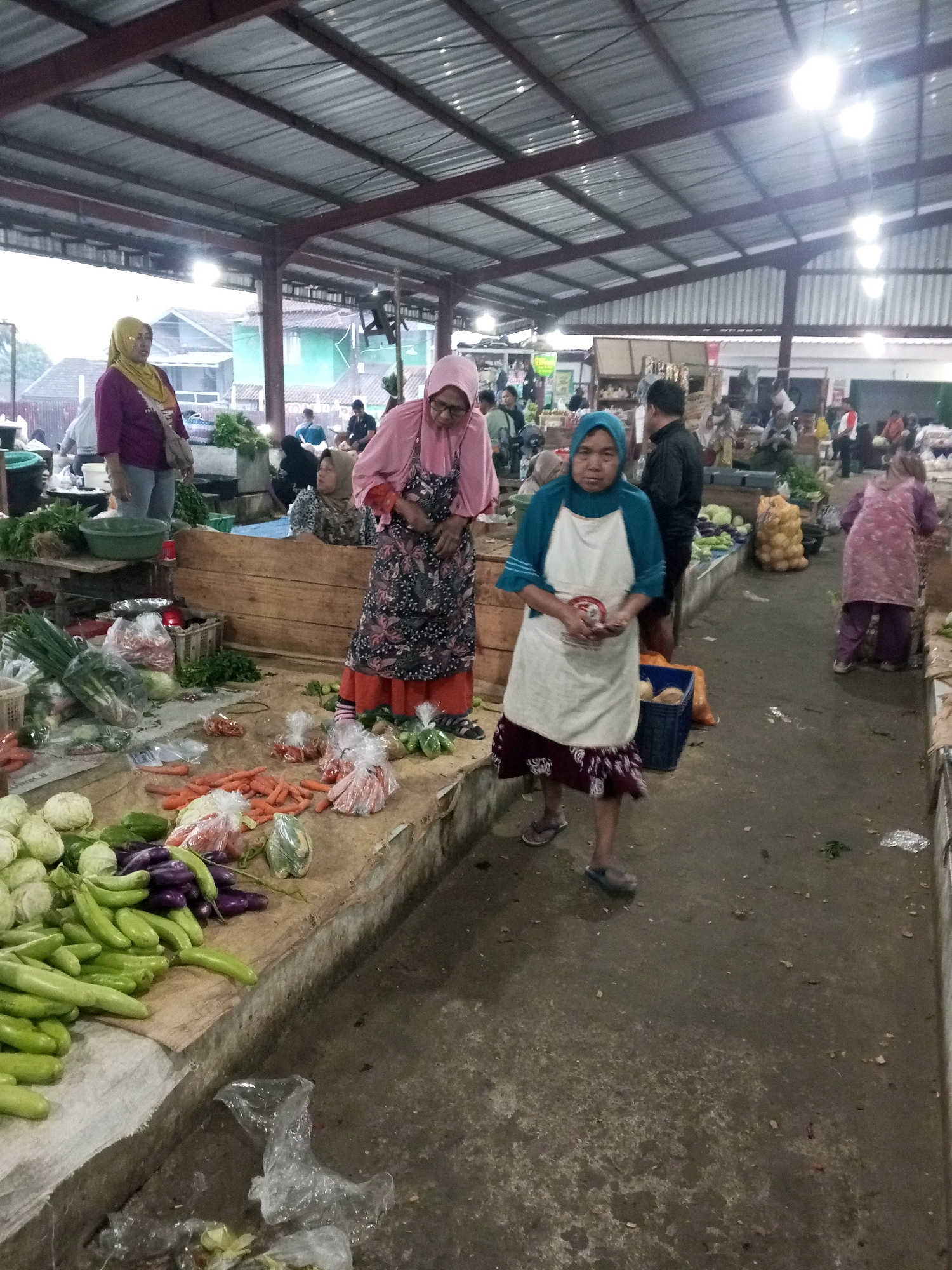 The image depicts an outdoor market scene in Gentan, Indonesia. A covered structure with a corrugated metal roof supported by metal beams provides shelter. In the foreground, a woman wearing a blue hijab and a white apron stands behind a stall laden with fresh vegetables, including green beans, eggplant, cabbage, and carrots. She is holding what appears to be a small item in her hands. To her left, another woman in a pink hijab and a patterned dress stands near a display of vegetables. In the background, various other individuals are visible, some tending to stalls or browsing. The ground is paved with concrete and shows signs of being a high-traffic area. The lighting suggests it might be early morning or late afternoon.