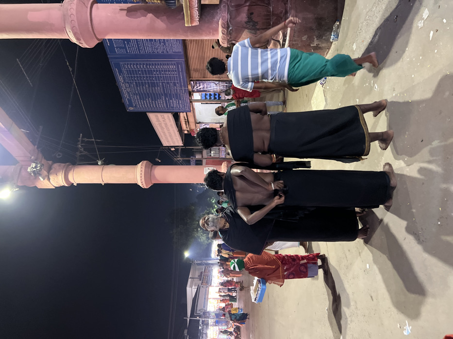 The image depicts a bustling outdoor scene at night, likely near a temple or pilgrimage site in Palani, India. Several people are visible, illuminated by artificial lights against a dark sky.
In the foreground and midground, a group of men stands, some shirtless and wearing traditional black dhotis or lungis. This attire is characteristic of Ayyappan devotees observing a vratham (vow) for their pilgrimage. One man, further back, wears a striped white and blue shirt with green pants. Another person, with a long white beard, is seen wearing dark clothing. In the distant background, more people are visible, some gathered around stalls or shops.
The scene is dominated by large, light-colored (possibly pink or beige) architectural pillars or columns, suggesting a temple entrance or a grand gateway. Overhead, electrical wires and light fixtures are visible. The ground is a light-colored paved surface, and long shadows are cast by the people, indicating strong light sources from the side.
Notable details include a large blue information board with columns of text, mostly illegible. Below it, a smaller white sign clearly shows the words "SABARIMALA" and "AYYAPPAN KEEPING PLACE," which strongly confirms the presence of devotees undertaking the Ayyappan pilgrimage. The overall atmosphere suggests a religious gathering or a transit point for pilgrims.