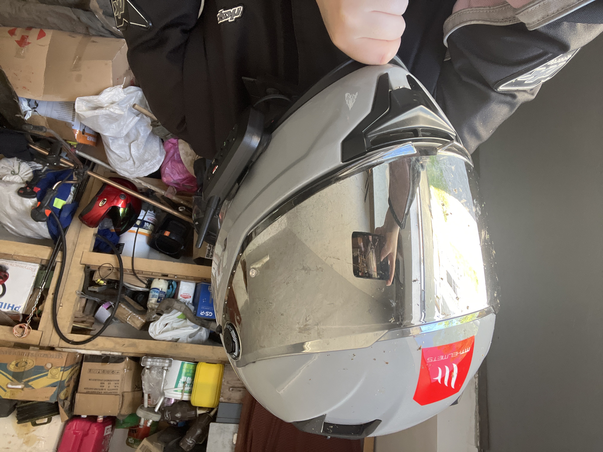 Here's a detailed description of the image:
**Scene:** The image appears to be taken in a cluttered garage or storage area. Various objects are visible in the background, including boxes, containers, bottles, tools, and what looks like another helmet.
**Subject:** The primary subject is a gray and silver motorcycle helmet held up by a person wearing a black and gray jacket. The helmet has a reflective visor and a red sticker with the brand "MT Helmets". The visor is dirty with smudges and bugs.
**People:** A hand and part of a jacket are visible, suggesting a person holding the helmet. The reflection in the visor of the helmet shows the image being taken on a cell phone.
**Environment:** The background is filled with clutter, indicating a possibly disorganized storage space.
**Location:** The location is not determinable from the image. Cherliany, Ukraine is not determinable from the image.