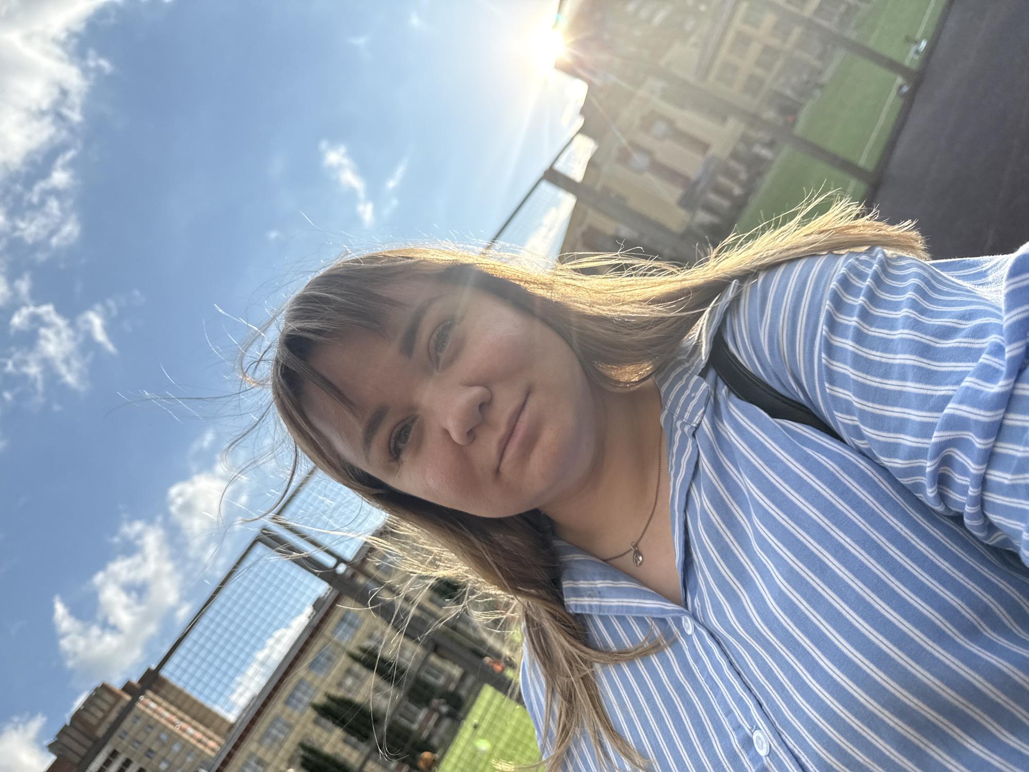 The media file is a selfie taken outdoors on a sunny day. The subject is a young woman with light brown hair, wearing a light blue and white striped button-up shirt and a delicate silver necklace. She is looking directly at the camera with a neutral expression.
The background features a bright blue sky with scattered white clouds. The sun is high in the sky, casting lens flares and illuminating the scene. Behind her, a chain-link fence runs across the frame, and beyond that, buildings with windows and a green sports field are visible. The architectural style of the buildings suggests an urban or institutional setting.
The overall impression is of a casual, daytime photo, possibly taken during a break or outing. The sunlight and clear sky indicate pleasant weather. The presence of the sports field and fenced area might suggest a park, school, or recreational facility. There is no visible text in the image.