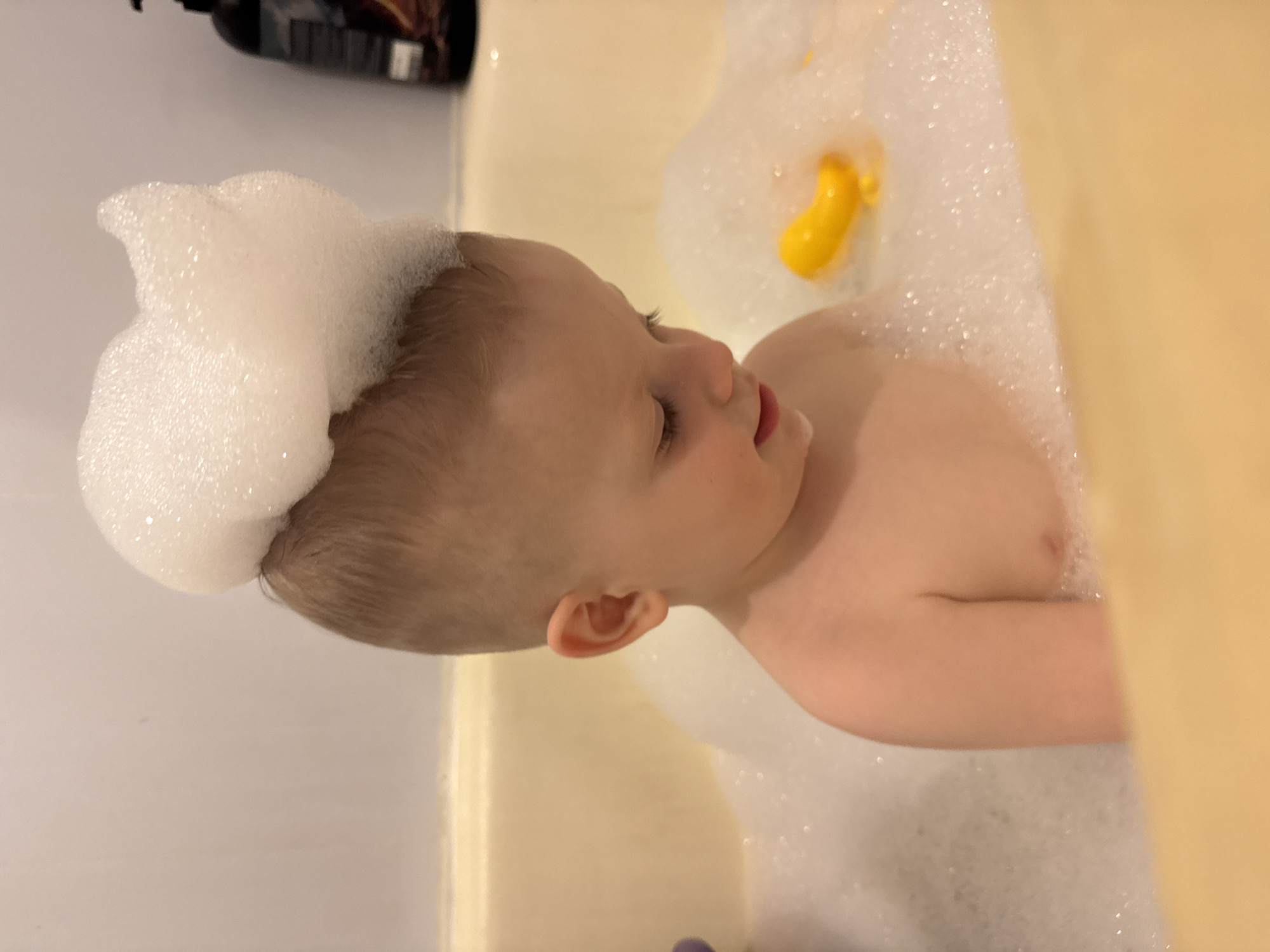 An infant is seen in a bathtub filled with white, foamy bubbles, indicating a bubble bath. The child, with light hair and fair skin, is partially submerged in the water, facing towards the right side of the frame with a slightly open mouth. A substantial amount of white foam sits atop the baby's head like a cap. Their right ear is clearly visible.
In the bubbly water, a bright yellow bath toy, possibly a duck or a similar animal, can be seen floating. The bathtub itself appears white or light cream in color. In the blurred background, a dark bottle, likely shampoo or body wash, is visible. The scene suggests an indoor setting, likely a bathroom, given the context of a bath. The overall atmosphere appears calm and playful. The location is Saint Petersburg, Russia. No discernible text is visible on any objects.