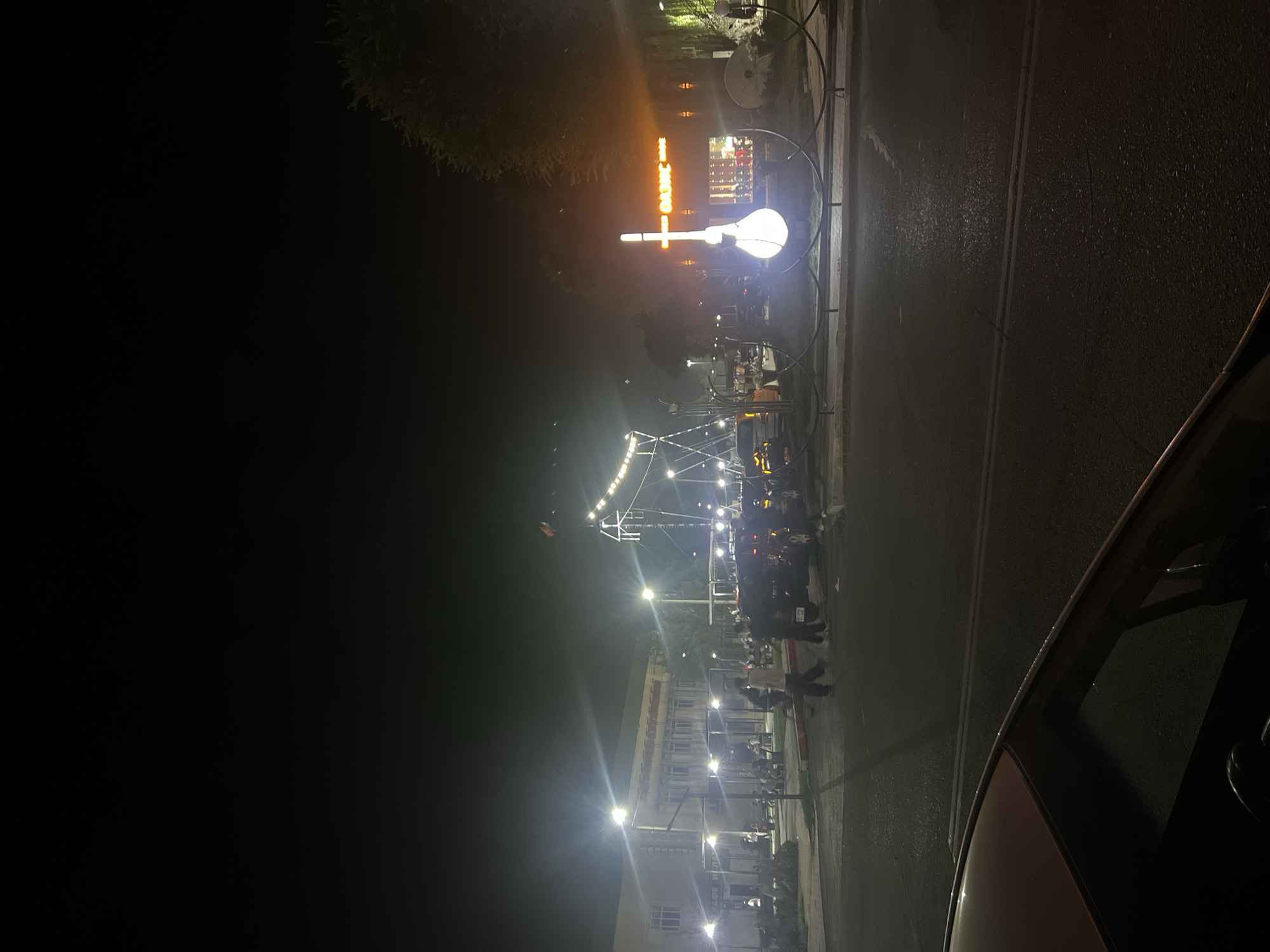 The image captures a nighttime street scene in Ak-Suu, Kyrgyzstan, near a building that appears to be a cafe or restaurant, indicated by signage that includes Cyrillic text and what looks like "КАФЕ ИЛА" or similar. Several people are visible, some walking on the sidewalk and street, while others are seated at tables outside the building. Streetlights illuminate the area. To the right of the frame, there's a prominent illuminated sculpture shaped like a musical instrument, possibly a lute or a similar stringed instrument, set against a backdrop of trees and a building with orange neon signage that reads "OASIS". Cars are parked and driving on the street. In the foreground, the hood and windshield of a car are visible, suggesting the photograph was taken from within a vehicle. The overall atmosphere is lively, with a mix of pedestrian and vehicular activity under artificial lighting.