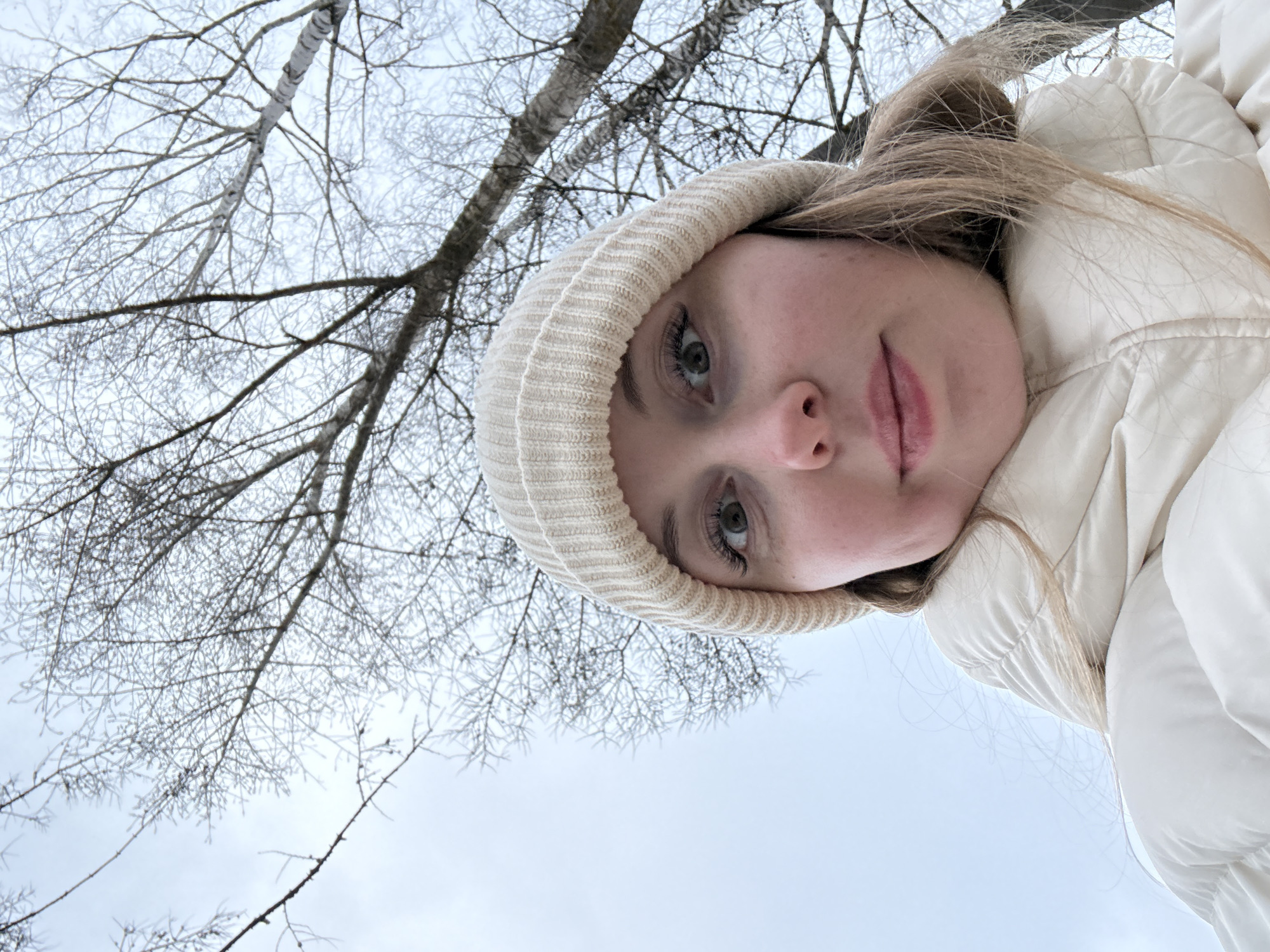 A low-angle selfie of a woman wearing a light-colored beanie and puffer jacket. The background features bare tree branches against a pale sky, possibly indicating winter or early spring. The location is Unknown, Unknown.