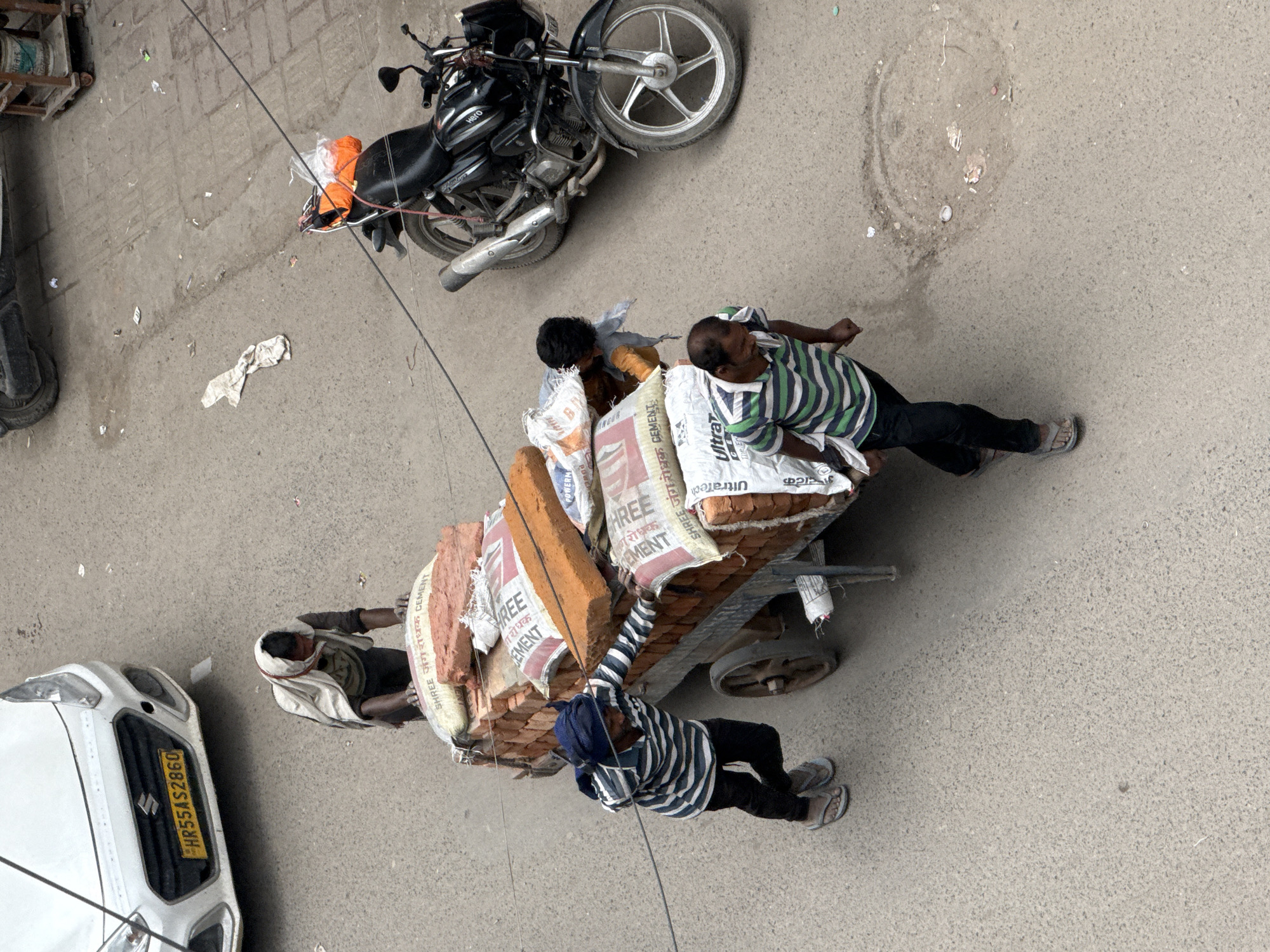 The overhead shot captures a bustling street scene in Delhi, India, where several individuals are engaged in manual labor. A white car with a yellow license plate bearing the registration "HR55AS2860" is parked on the left side of the frame. In the center, a two-wheeled cart is laden with a substantial load of bricks and sacks of cement. Three men are actively involved in moving this cart. One man, wearing a white cloth draped over his head and shoulders, is pushing the cart from behind. Another man, dressed in a blue and white striped shirt and dark pants, is positioned on the left side of the cart, also applying force to move it. A third individual, wearing a green and white striped shirt and dark pants, is on the right side of the cart, appearing to be pushing or guiding it. A fourth person, partially obscured by the cart and the other individuals, is visible on the right side, seeming to assist with the load. The street is paved with asphalt and shows signs of wear and tear, including a circular manhole cover. A black motorcycle with an orange bag strapped to its rear is parked on the right side of the image, facing away from the viewer. The environment appears to be an urban setting, with scattered debris on the ground, typical of a busy street in India. The lighting suggests it is daytime.