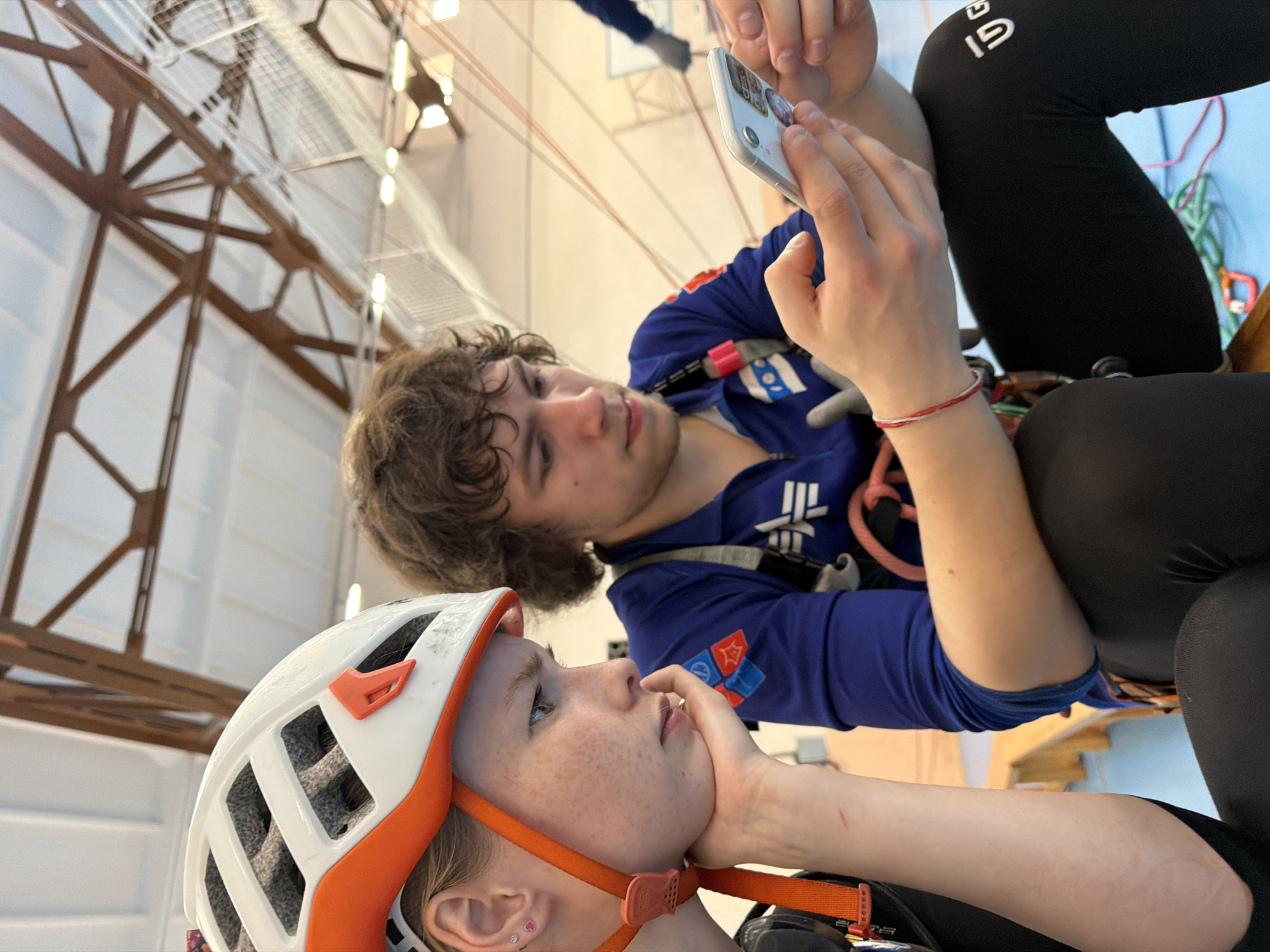 Two individuals are suspended, presumably secured with climbing gear, in what appears to be an indoor climbing facility. A female wearing a white and orange climbing helmet, with freckles visible on her face, rests her chin on her hand. Behind her, a male, also wearing climbing gear, is focused on a smartphone. The environment features visible structural steel framework near the ceiling, and portions of a climbing wall are visible. The image suggests a moment of rest or preparation before or after climbing, possibly at Berezniki, Russia.