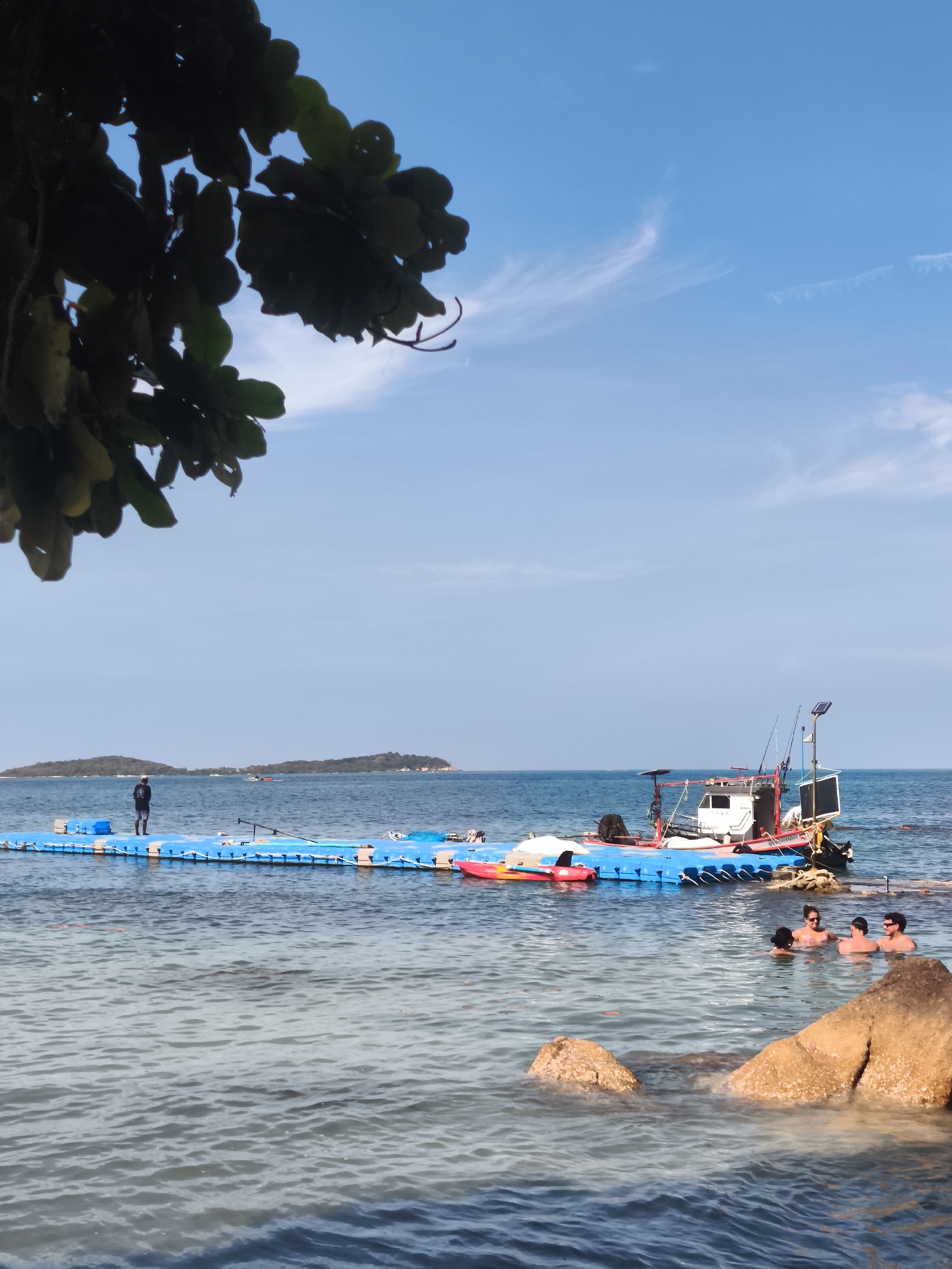 The image depicts a coastal scene with a floating pontoon walkway extending from the shore into the ocean. A person stands on the pontoon, facing away from the camera, looking towards two islands in the distance. A small boat is moored to the pontoon. In the foreground, several individuals are swimming and wading in the clear, shallow water. The scene is set against a backdrop of a blue sky with scattered white clouds. Rocks are visible both in the water and partially submerged. The location appears to be a beach or a resort area in Ko Samui District, Thailand.