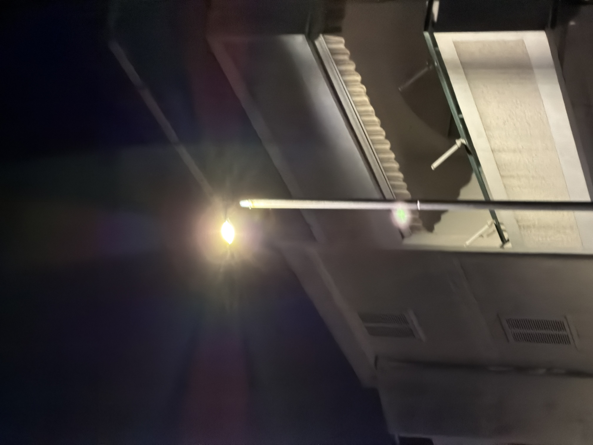 An outdoor, night-time, eye-level, medium shot shows a modern streetlamp with a bright white light illuminating the dark sky. The streetlamp is positioned to the left of the frame, with its pole extending vertically upwards. In the mid-ground, a building facade is visible, featuring horizontal lines and what appear to be louvered windows. The building's architecture suggests it could be an apartment complex or a residential building. A balcony or overhang with a striped awning is visible on the upper portion of the building. The lighting from the streetlamp creates lens flares and color aberrations in the dark sky. The scene is devoid of any people or discernible activity, presenting a static, architectural view. The environment suggests an urban setting, likely in Budva, Montenegro, given the architectural style and the presence of street lighting.