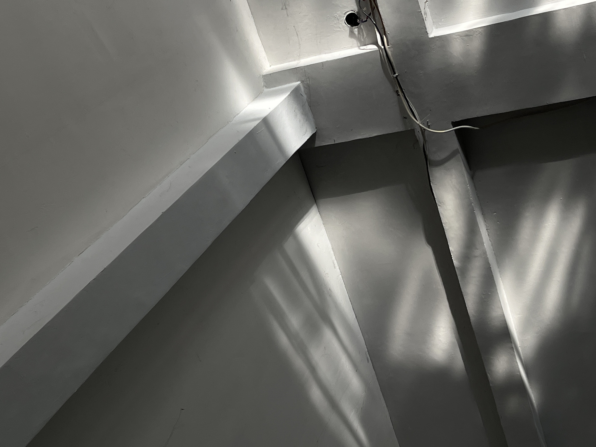 The image shows a close-up, low-angle interior view of a building under construction or renovation in Kedawung, Indonesia. The scene is dominated by concrete beams and walls, rendered in shades of gray and white. Sunlight streams in from an unseen source, casting long, parallel shadows that create a dramatic, striped pattern across the surfaces. These shadows suggest that the light is originating from a barred window or external structure.
Several structural elements are visible, including a prominent horizontal beam running diagonally across the upper portion of the frame, a smaller beam or ledge below it, and vertical corners where walls meet. The surfaces appear unfinished, with visible texture and some imperfections. Electrical conduits and wires are present along one of the walls, with a circular opening suggesting an electrical junction box or outlet. The overall impression is one of a utilitarian, possibly industrial or unfinished space, illuminated by natural light.