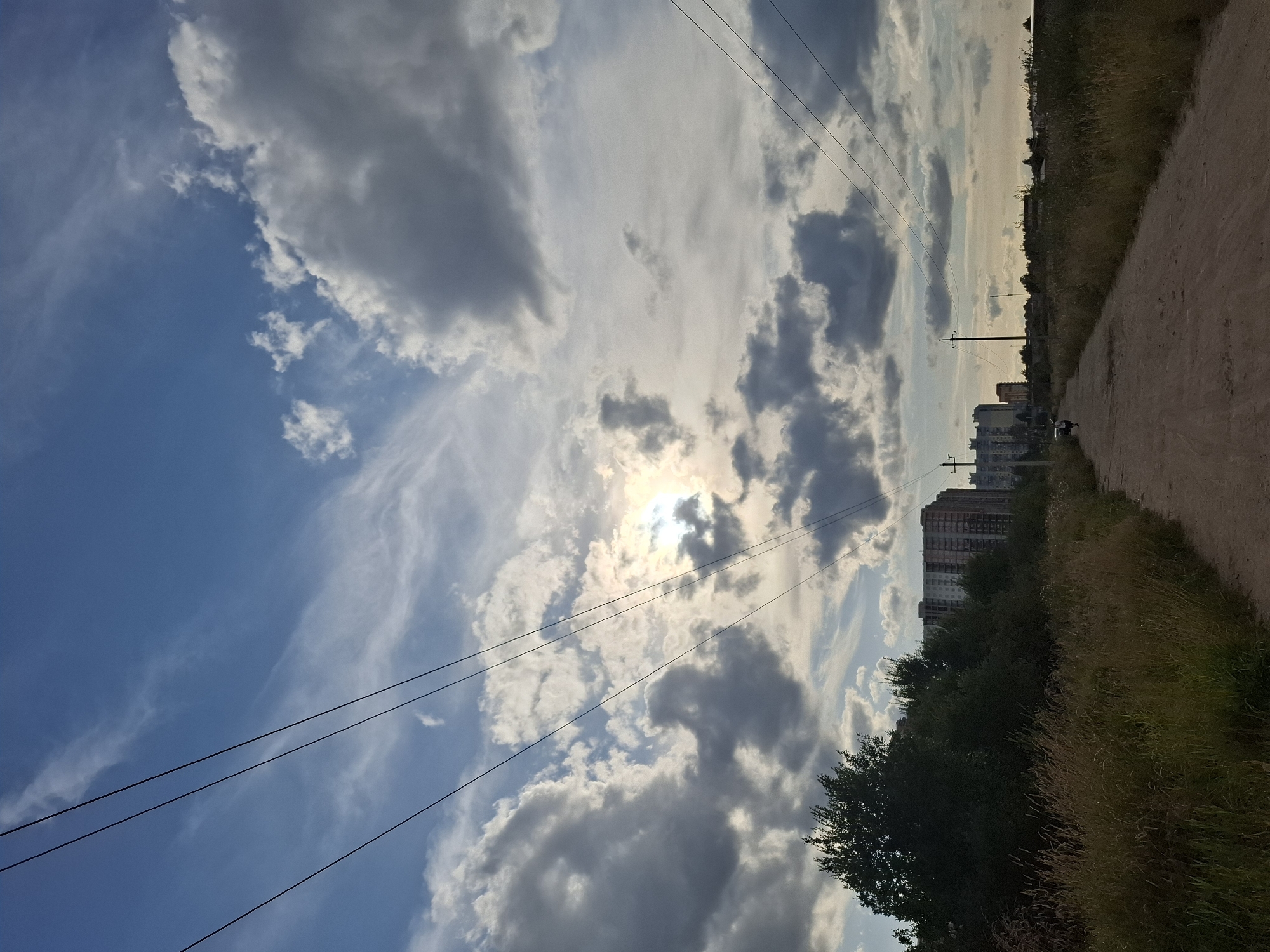 The image depicts a rural road leading towards a city skyline. The road is unpaved and appears to be made of dirt or gravel. On the left side of the road, there is tall, dry grass and a dense line of trees. Beyond the trees, several multi-story apartment buildings are visible, characteristic of urban development. On the right side of the road, there is also tall grass, and in the distance, more buildings can be seen. Overhead, power lines stretch across the frame, supported by utility poles. The sky is filled with a dramatic mix of clouds, with patches of blue sky and the sun peeking through. There is a single person on a bicycle riding down the road in the distance. The overall scene suggests a transition between a rural and urban environment, possibly on the outskirts of a city. The presence of apartment buildings and power lines indicates an inhabited area, while the unpaved road and natural vegetation suggest proximity to undeveloped land. The lighting suggests it is either early morning or late afternoon.