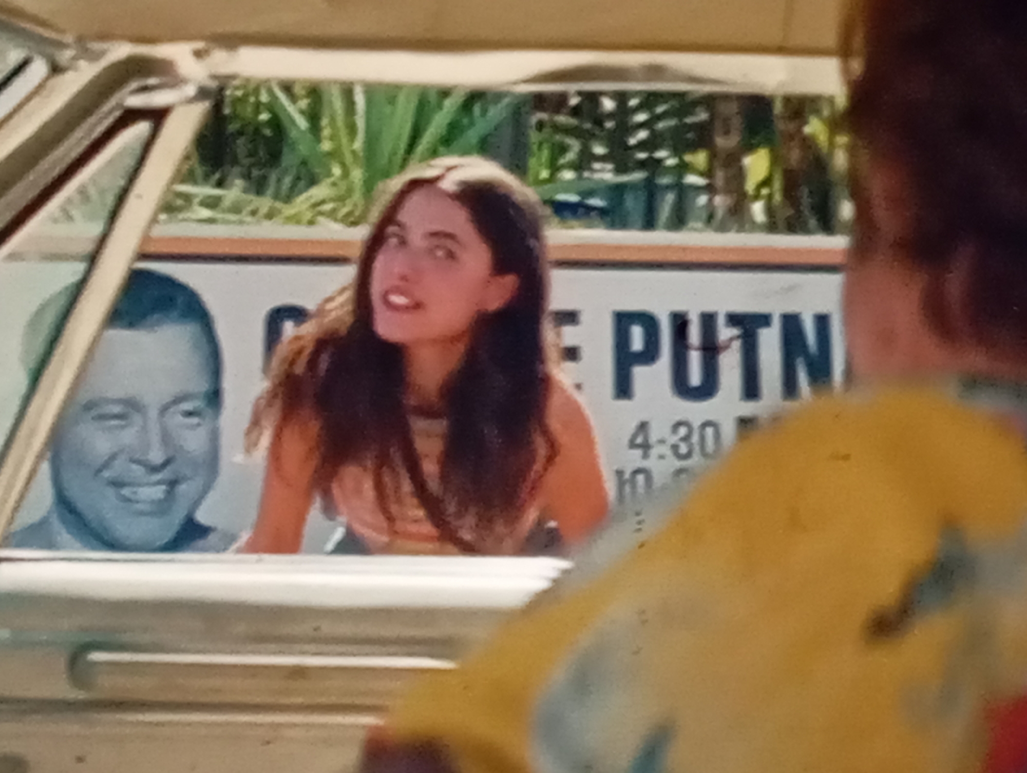 The image captures a close-up view of people inside and outside a vehicle, with a sign in the background. In the foreground, slightly out of focus, is the back of a person wearing a yellow shirt with a tie-dye pattern.
Through the car window, a young woman with long, dark hair is visible. She is looking towards the right of the frame with a surprised or excited expression, her mouth open. She is wearing a light-colored, possibly pink, top. To her left, a portrait of a smiling man is displayed on a sign, rendered in blue tones. The man appears to be middle-aged, with fair skin and dark hair.
Behind the people, a sign with text is partially visible. The text includes "PUTN" in large, dark letters, and below that, "4:30" and what appears to be "10." The background behind the sign shows lush green foliage, suggesting an outdoor setting, possibly with trees or large plants. The lighting in the image is bright, indicating it was likely taken during the daytime. The overall impression is a candid moment captured, perhaps at a roadside stop or event.