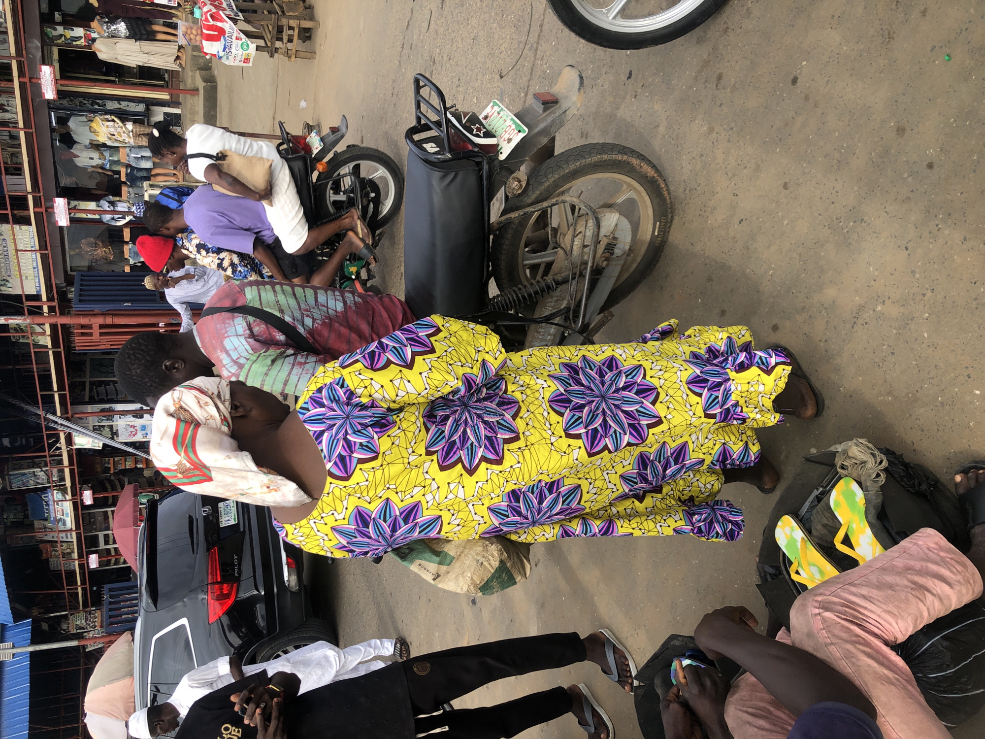 A street scene in Ikeja, Nigeria features a woman in a bright yellow and purple floral dress standing in the foreground, her back mostly to the camera. She wears a white headscarf with a patterned band. To her left, a man in a black t-shirt and black pants with white drawstrings is visible from the waist down, wearing grey flip-flops. He appears to be interacting with a dark bag containing colorful yellow and green flip-flops. To the left of the woman, a black Honda Pilot SUV is parked. Behind the woman, a motorcycle is parked, with three people on it: a man in a purple shirt, a man in a patterned shirt with a red cap, and a woman in a white top. Several market stalls with clothing displays are visible in the background. The ground is paved and appears to be dusty.