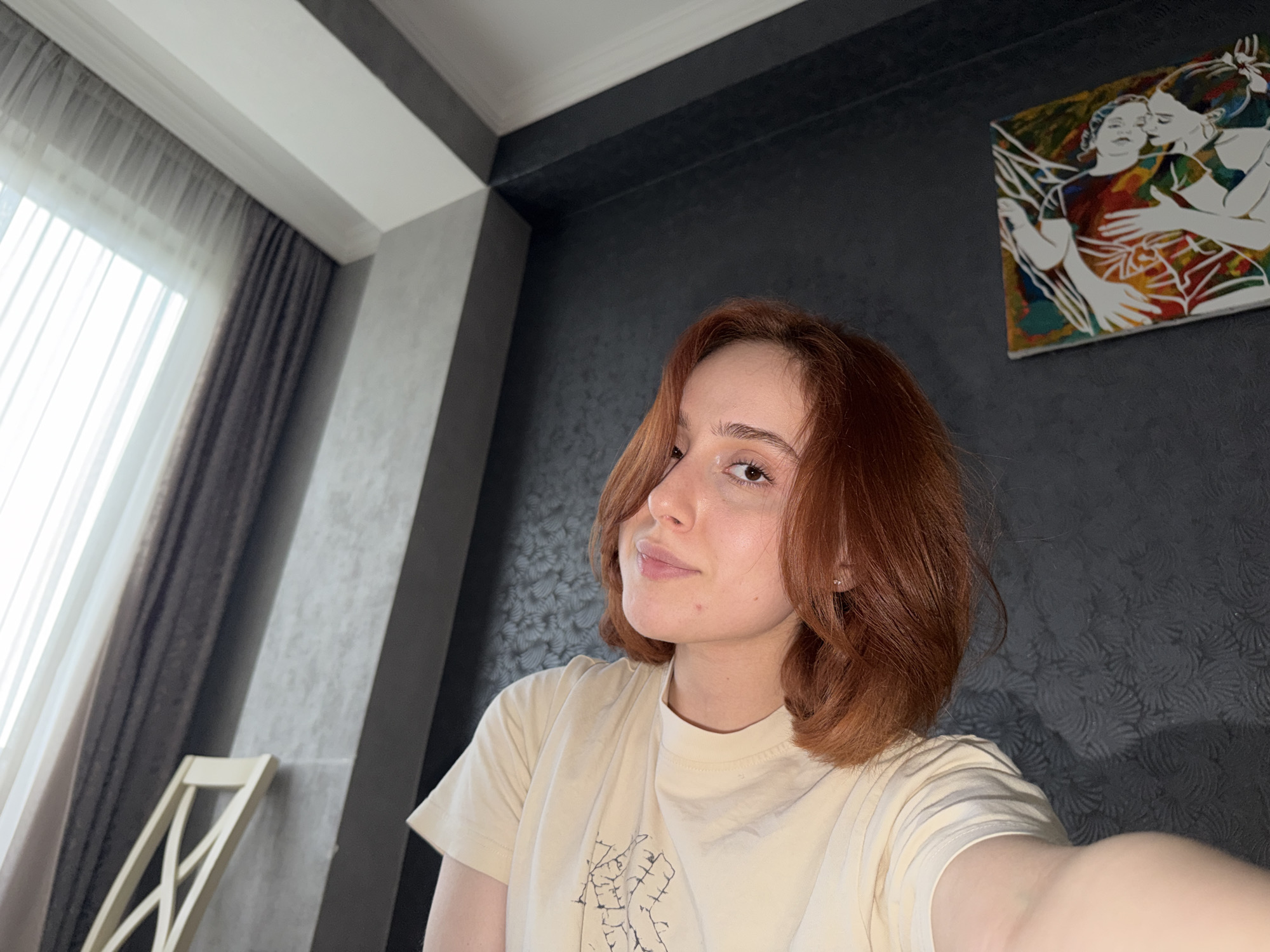 A young, fair-skinned woman with auburn bobbed hair and a slight smile is taking a selfie indoors. She is wearing a light-colored t-shirt with a faint design. Behind her is a dark, patterned wall with an abstract painting depicting two embracing figures with multi-colored accents. To the left, a window with a light curtain and a darker drape is visible. A wooden chair is partially visible. The location may be Tbilisi, Georgia, given the architectural style and patterns.