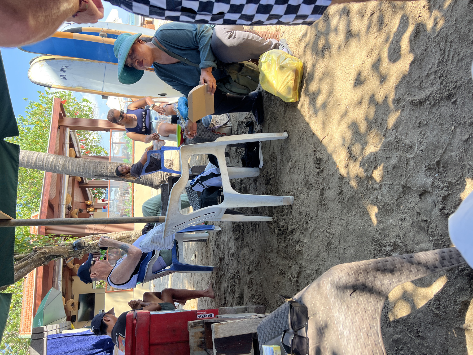 The image depicts a scene on a beach in Kuta, Indonesia, likely near a beachside cafe or establishment. The ground is covered in sand, with some shadows cast by overhead foliage and structures.
In the foreground, a person wearing checkered shorts is partially visible on the right. To the left, a woven plastic chair sits on the sand, with a pair of sunglasses resting on a textured surface in front of it.
Further into the scene, several people are seated. On the left, a man wearing a blue cap and sunglasses is sitting in a chair, holding a bottle to his mouth. Next to him, another person in a black t-shirt and shorts is also seated.
In the middle ground, an older man in a white tank top and a blue baseball cap is seated, holding a bottle. Beside him, a white plastic chair is empty. A woman with a blue bandana and sunglasses is seated, wearing a dark blue tank top with the word "STRONGER" on it. Another person is seated behind her.
On the right side of the middle ground, a woman wearing a teal top and a wide-brimmed teal hat is seated, holding a beige takeout container and looking to her right. A green bag is on the sand beside her, along with a bright yellow plastic bag.
In the background, three surfboards are leaning against a structure. A beach umbrella provides shade over some of the seated individuals. A building with a tiled roof and open-air seating is visible in the distance, suggesting a commercial area along the beach. The overall atmosphere appears relaxed, with people enjoying their time outdoors.