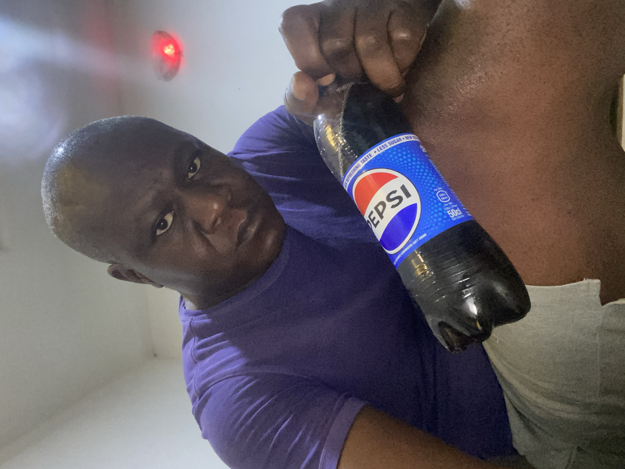 A medium-shot photograph shows a dark-skinned man wearing a purple t-shirt, holding a plastic bottle of Pepsi. The camera angle is from below, looking upwards at the subject. A red light is visible on the white ceiling. The lower part of the person’s leg, wearing grey shorts, is also visible. There is no information to confirm the location in Kubwa, Nigeria.