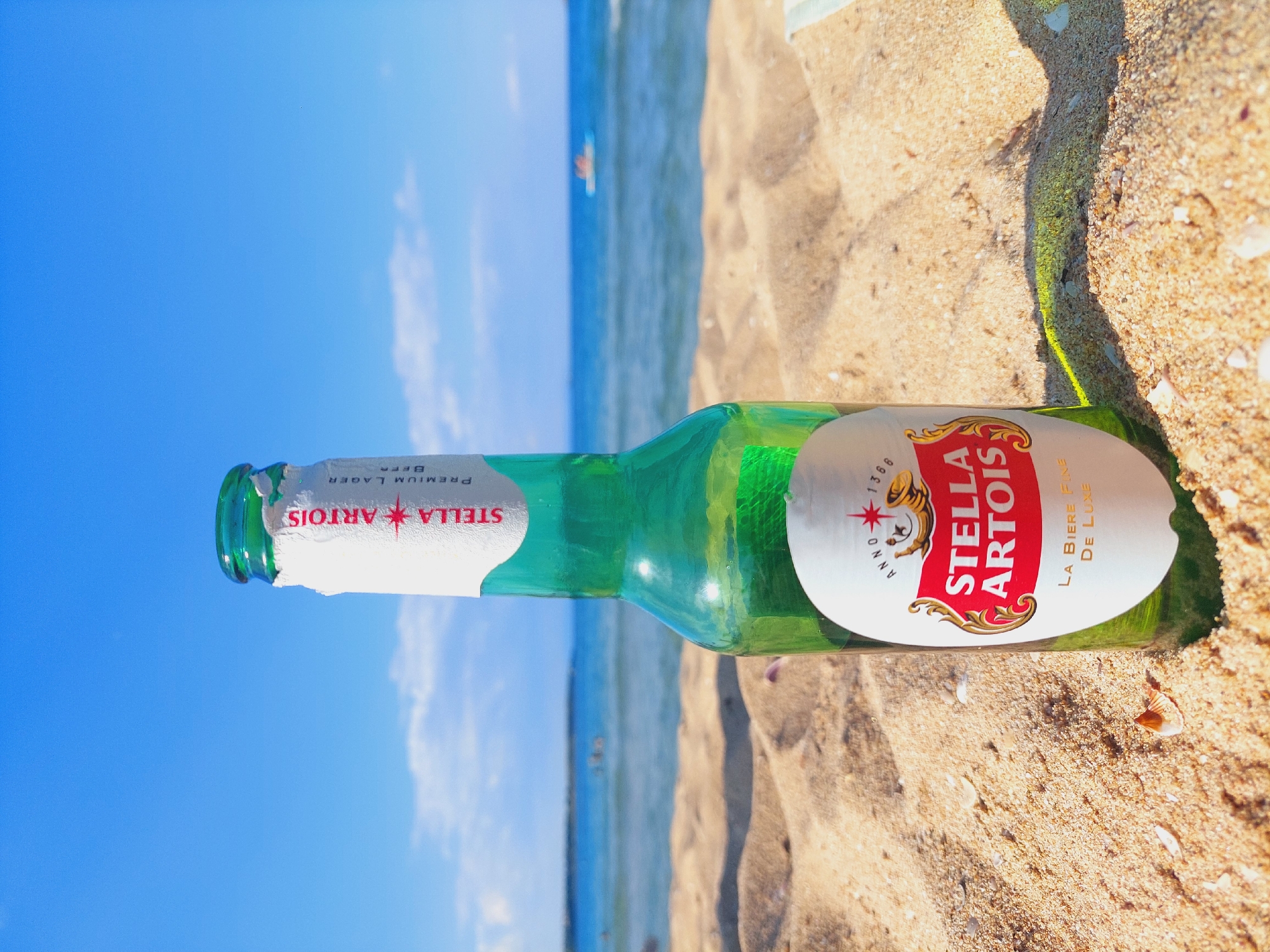 A green glass bottle of Stella Artois Premium Lager beer lies on its side in the sand on a beach. The bottle is mostly intact, with a white label that has red and gold accents. Text on the label reads "STELLA ARTOIS" and "PREMIUM LAGER BEER". Another section of the label has text that reads "ANNO 1366" and "LA BIÈRE FINE DE LUXE".
The background shows a bright blue sky with some wispy white clouds and the ocean. The water is a clear blue and appears calm, with a few distant figures visible in the water, suggesting people are swimming or enjoying the sea. The sand is light brown and looks dry and granular, with a few small seashells visible. The sunlight appears bright, indicating it's likely daytime, possibly mid-morning or afternoon, on a sunny day. The scene evokes a relaxed, leisurely atmosphere associated with a beach vacation. The location context is Sozopol, Bulgaria.