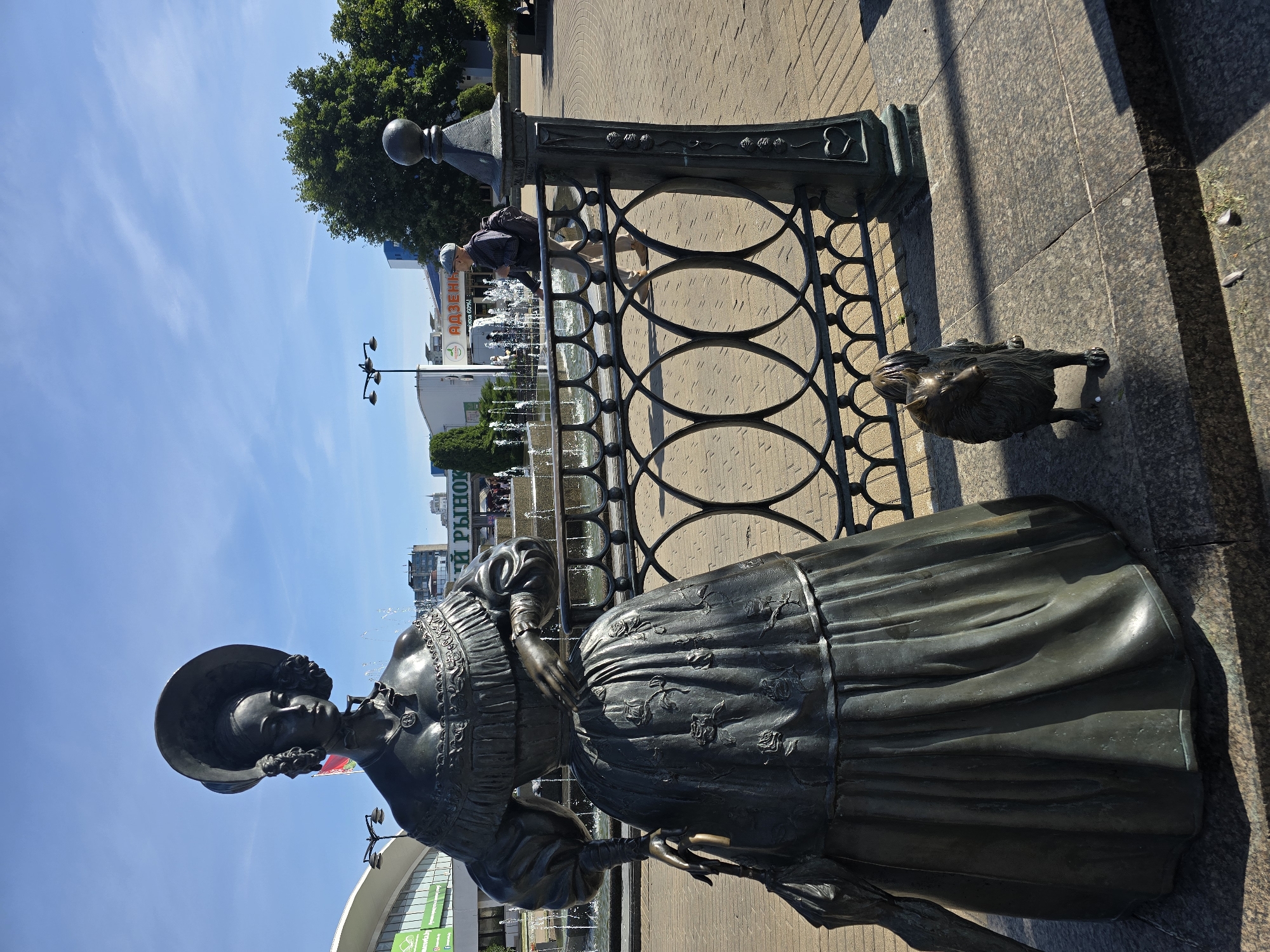 A bronze sculpture of a woman in a voluminous dress and wide-brimmed hat stands to the left of a black metal fence. A smaller bronze sculpture of a dog is positioned in front of the fence to the right of the woman. In the background, a man wearing a cap, dark jacket, and light pants walks away from the camera, past a fountain with multiple jets of water. Beyond the fountain, there are several buildings and trees. The scene is set in a paved outdoor plaza on a sunny day with a clear blue sky. The sculpture of the woman and dog is located in Minsk, Belarus, specifically in front of the "TsUM" department store.