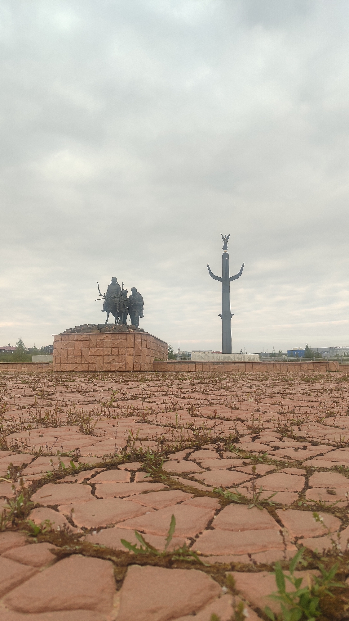 The image shows a monument in the city of Mirny, Russia, on an overcast day. In the foreground, a paved area with red interlocking bricks is covered with sparse green vegetation.
The main monument, located in the midground, features a bronze sculpture of three figures, two standing and one on horseback, dressed in what appears to be historical or traditional clothing. They are positioned on a tiered, rock-faced pedestal.
To the right of the sculptural monument, a tall, dark, slender obelisk-like structure rises from the ground. This obelisk is topped with a winged figure, possibly an angel or a bird. The background reveals a low-rise cityscape with buildings and trees under a cloudy sky. The overall impression is of a civic memorial or monument.