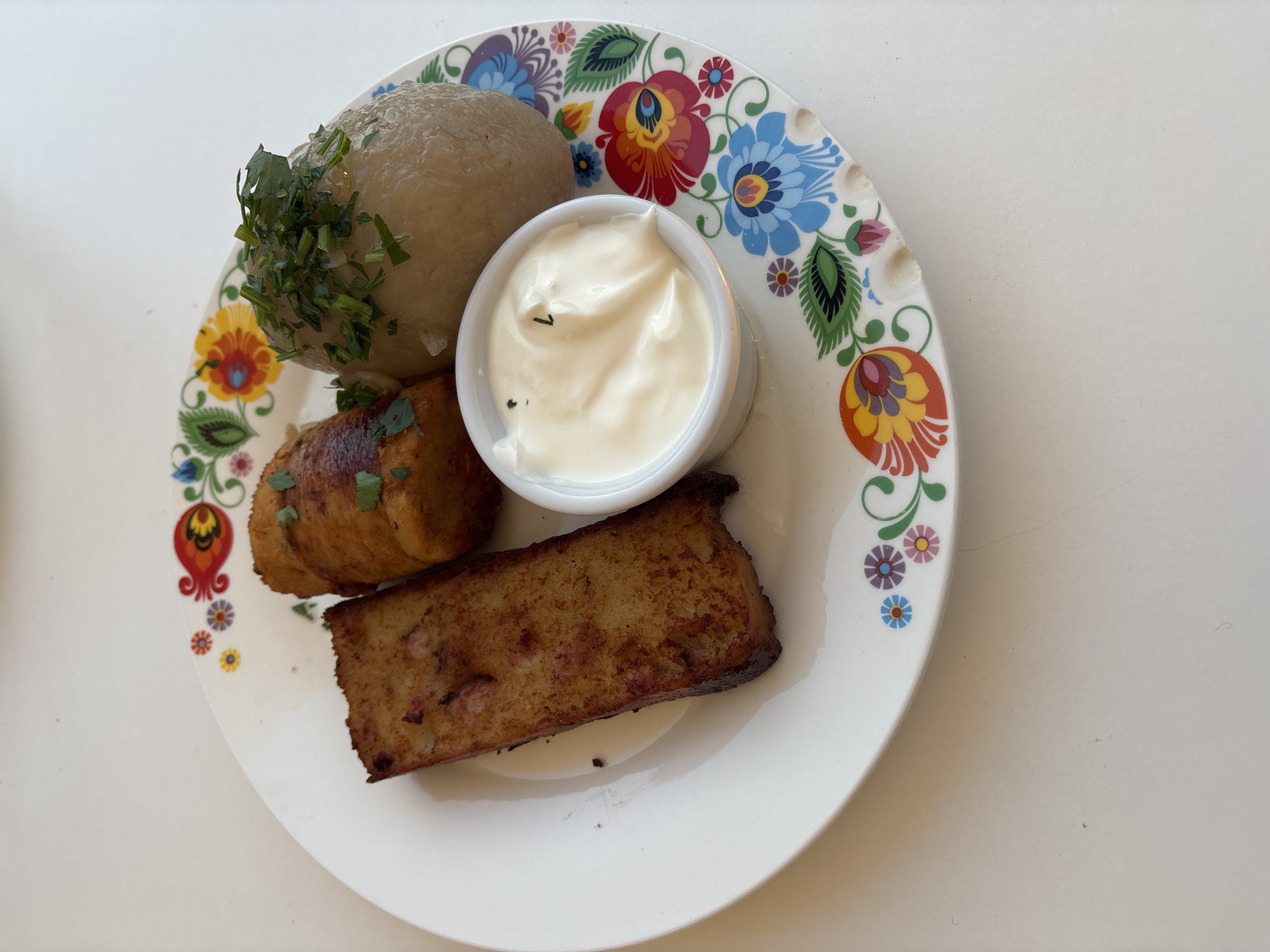 A close-up, overhead shot captures a white plate adorned with colorful folk-art style floral patterns, filled with a meal. The plate contains three food items and a small dish of white sauce.
On the left is a rectangular piece of what appears to be potato pancake, golden brown and slightly crispy, with visible pieces of red and white mixed in, suggesting it might contain meat or fat.
In the center, a single, oblong potato dumpling, referred to as a "kartacz" or "cepelinai" in Lithuanian cuisine, sits on the plate. It is roughly shaped and topped with finely chopped green herbs. This potato dumpling is a staple in Eastern European cuisine, particularly in Lithuania and Belarus, and is commonly served in regions with historical ties to these cultures, such as the Podlaskie Voivodeship in Poland, where Białystok is located.
To the right of the dumpling is a small, white ramekin filled with a creamy white sauce, likely sour cream or a similar dairy-based condiment, also garnished with a few specks of green herbs.
The plate rests on a light-colored, textured surface, possibly a tabletop. The lighting is bright and natural, casting soft shadows. The overall impression is of a hearty, traditional meal presented on a decorative plate.