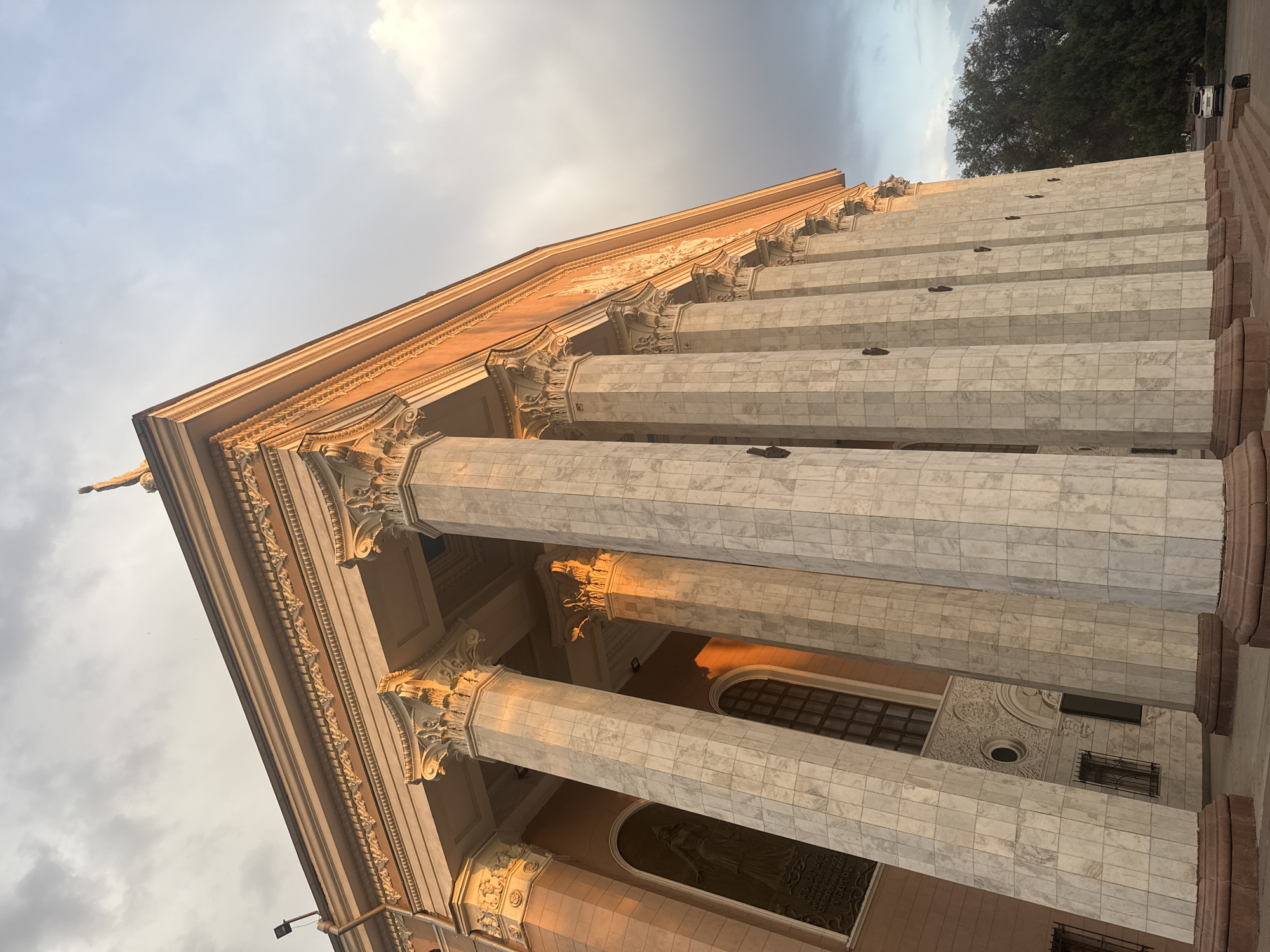 The image captures the front facade of the Kyrgyz National Academic Opera and Ballet Theatre in Bishkek, Kyrgyzstan, bathed in the warm glow of what appears to be either sunrise or sunset. The building is characterized by its imposing classical architecture, featuring a series of large, fluted marble columns topped with ornate Corinthian capitals. These columns support a prominent entablature adorned with decorative friezes. The facade itself is a pale ochre color, contrasting with the white of the marble.
To the left of the columns, a tall, arched window is visible, and below it, a large bronze relief plaque depicting a female figure. The overall impression is one of grandeur and historical significance. The sky above is partly cloudy, with a mix of soft grey and blue hues, suggesting dynamic weather conditions. A line of trees is visible to the right of the building, and a white car is parked in the distance, indicating the presence of a public space. The lighting creates long shadows, emphasizing the three-dimensional qualities of the architectural elements. The image is taken from a low angle, looking up towards the building, which enhances its monumental scale.