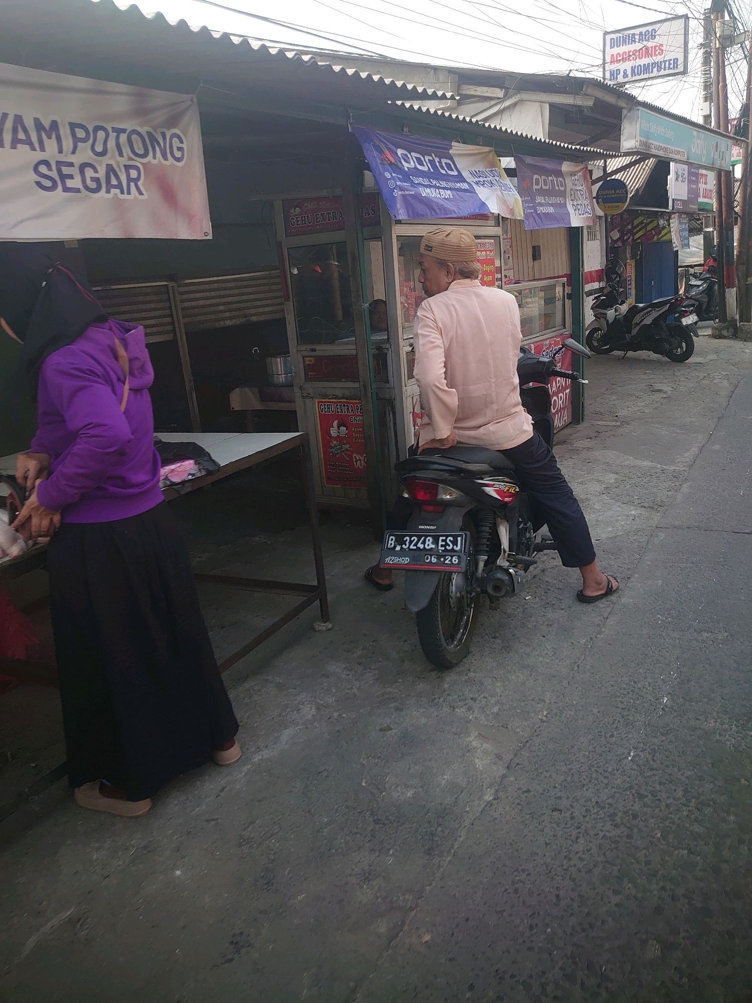 In Depok, Indonesia, on a street lined with small businesses, a person wearing a light pink shirt and dark pants sits on a motorcycle. They have a light-colored cap on their head and are facing away from the camera. To the left of the motorcycle, a person wearing a purple hoodie and a long black skirt stands next to a table where food items are being prepared, possibly chicken. Behind the table is a food stall with signage in Indonesian. In the background, more motorcycles are parked. The area appears to be a bustling street market or commercial area with awnings and various shop signs. One sign visible reads "DUNIA ACC AKSESORIES HP & KOMPUTER" indicating a shop selling accessories, mobile phones, and computers. Another sign advertises "AYAM POTONG SEGAR," meaning fresh cut chicken. The street is paved and shows signs of wear. The overall scene depicts a snapshot of daily life and commerce in an Indonesian city.