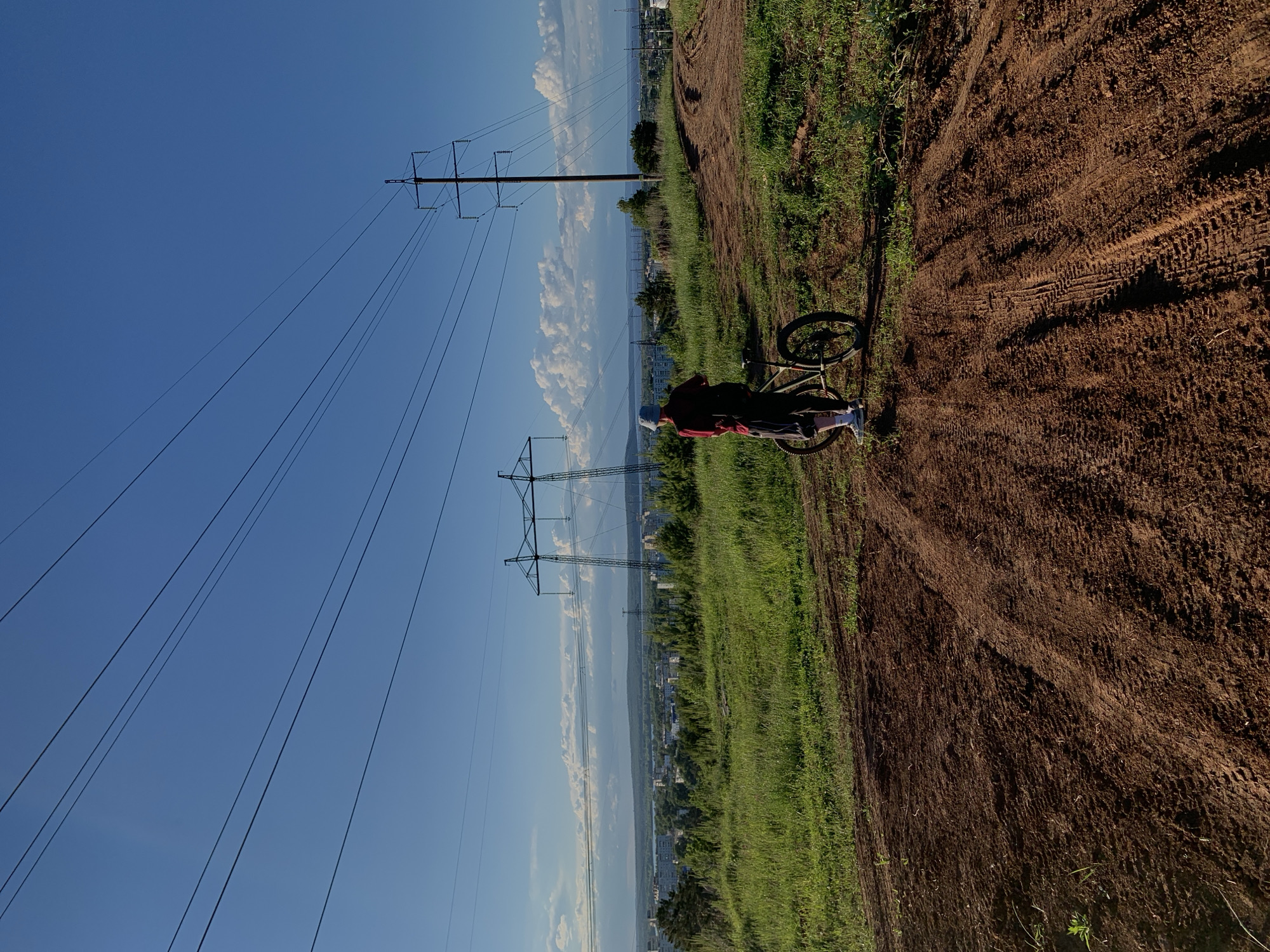 The image depicts a person on a bicycle, standing on a dirt path alongside a grassy area. The path shows tire tracks, indicating prior bike activity. Beyond the grass, there's a cityscape visible. Overhead power lines run parallel to the path. The sky is blue with some clouds. The location is identified as Chaikovsky, Russia.