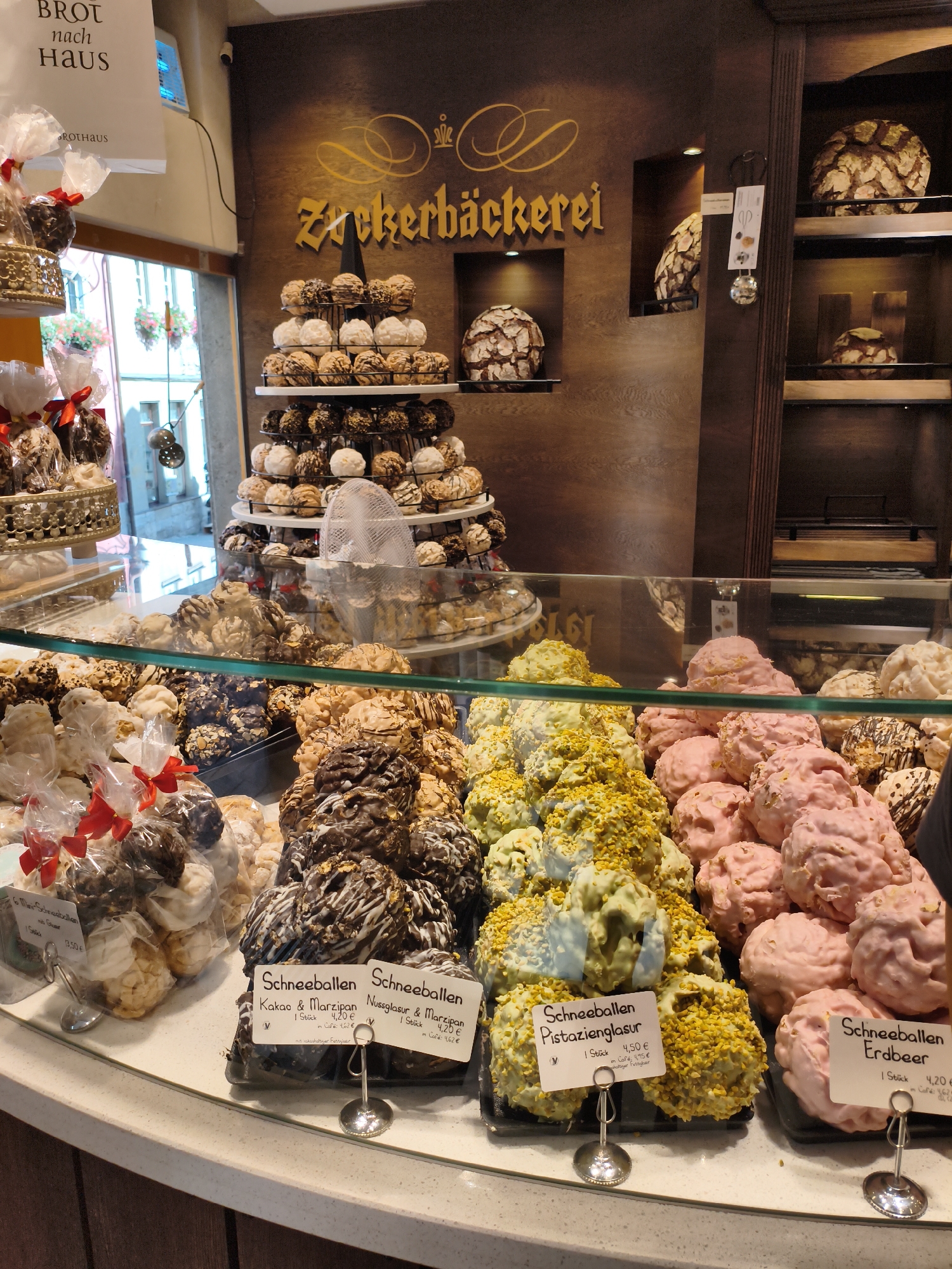 The image shows a display of confectionery in a bakery in Rothenburg ob der Tauber, Germany. The bakery's name, "Zuckerbäckerei," is visible on the wall in ornate golden lettering. The display case is filled with various round, pastry-like items. Some are covered in dark chocolate, some in white chocolate, some in what appears to be a light green or yellow icing with chopped nuts, and others are a light pink color. Several of these items are labeled with German text, including "Schneeballen," indicating a type of pastry.
In the background, on shelves, are several large, round loaves of bread with a textured, rustic crust. The overall environment appears to be a traditional German bakery, with wooden paneling on the walls and glass display cases showcasing the baked goods. The lighting is warm, creating an inviting atmosphere. There are no people clearly visible in the image, but the scene is set up for customers to browse and purchase items.
