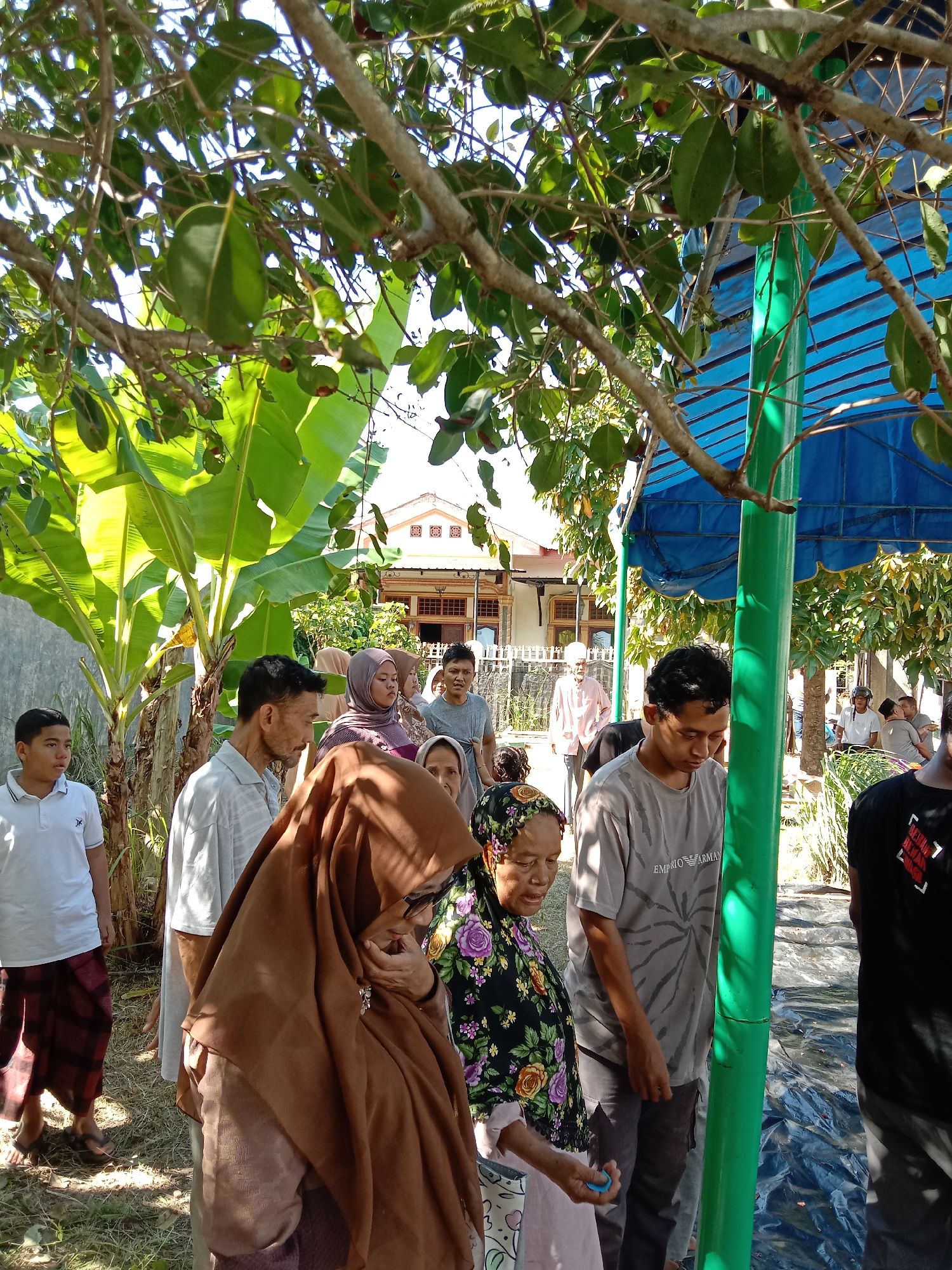 A gathering of people, including men, women and children, is taking place outdoors in Lhokseumawe, Indonesia. Some of the women are wearing hijabs. A banana tree is visible on the left. A house is visible in the background. A blue canopy is also visible, supported by green poles. A mix of overcast and sunny conditions is casting shadows on the scene. The ground appears to be covered with grass and some scattered debris.