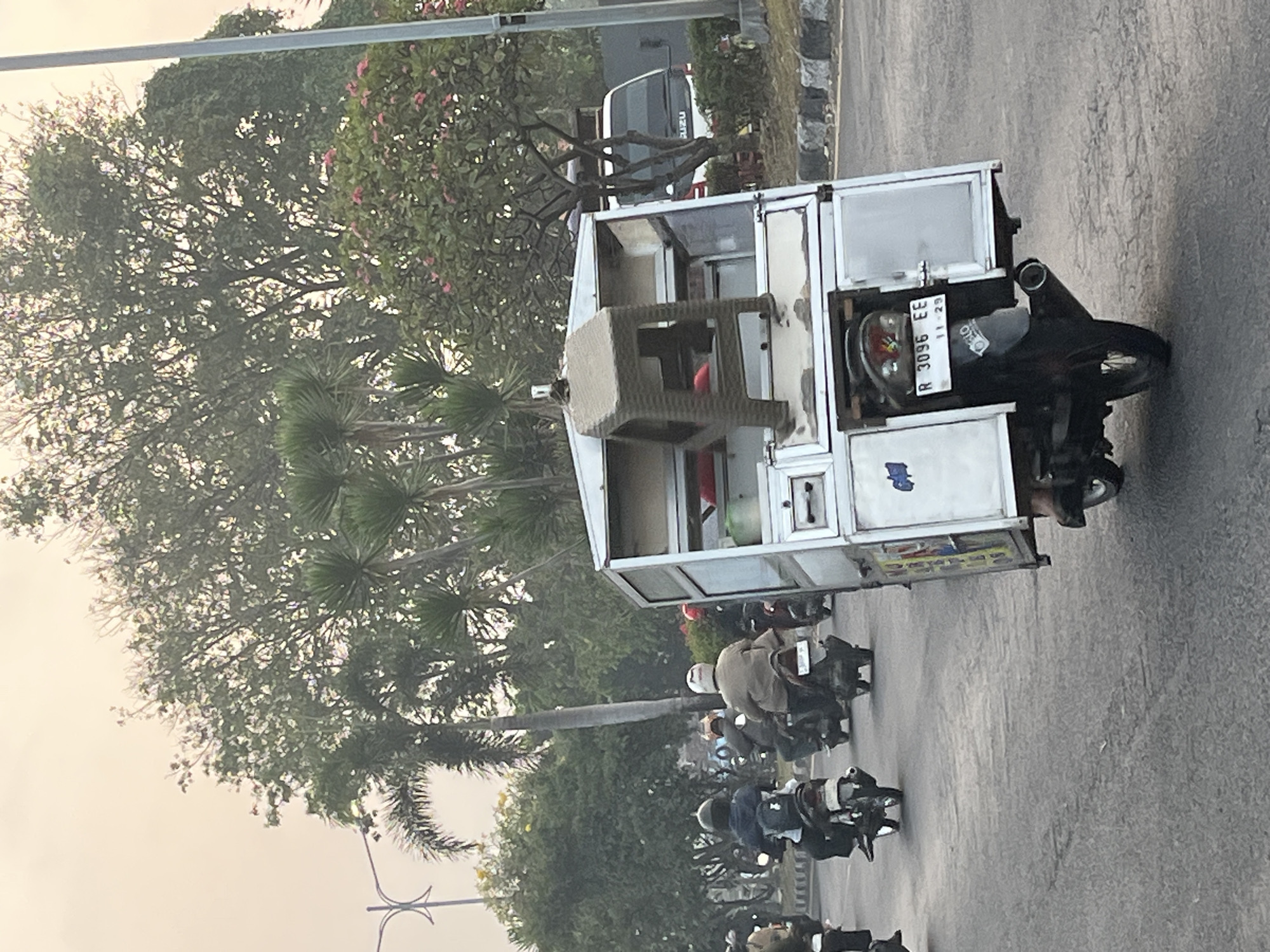 The image depicts a street scene in Kludan, Indonesia, with a modified motorcycle carrying a food cart traveling on a paved road. The motorcycle is black and is equipped with a white, box-like cart that appears to be made of metal. A plastic stool is secured to the top of the cart. The cart has a license plate that reads "R 3096 EE" with "11-29" below it. Several other motorcycles are visible on the road, some carrying passengers. The environment features lush green trees, including palm trees, and a hazy sky. In the background, a white truck with "ISUZU" visible on its front can be seen. The road has distinct lane markings, and a concrete barrier with stone aggregate lines the side of the road. The overall impression is of a typical day in a bustling Indonesian town.