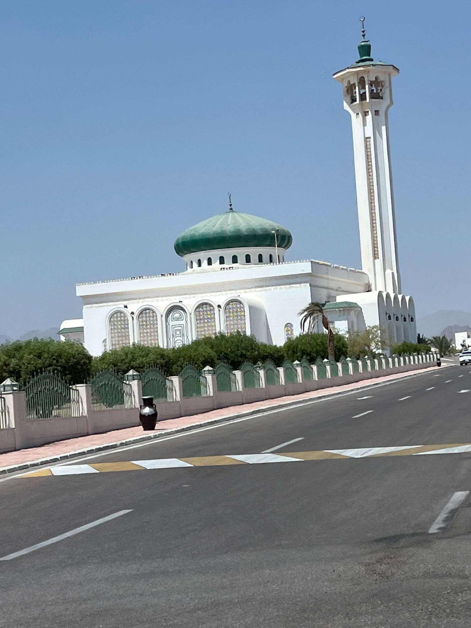 The image captures a daytime view of the Al Sahaba Mosque in Sharm El Sheikh, Egypt. The mosque features a prominent green dome and a tall, white minaret adorned with decorative golden bands and topped with a crescent moon finial. The main building of the mosque is white with arched windows featuring stained glass. A low green metal fence with decorative posts separates the mosque grounds from a paved road in the foreground. The road has clear white lane markings and a yellow and white speed bump. Lush green bushes and a single palm tree are visible behind the fence. In the distance, hazy mountains can be seen. A large dark vase stands on the sidewalk near the fence. A white vehicle is visible on the road in the far right of the image. The sky is clear and blue.