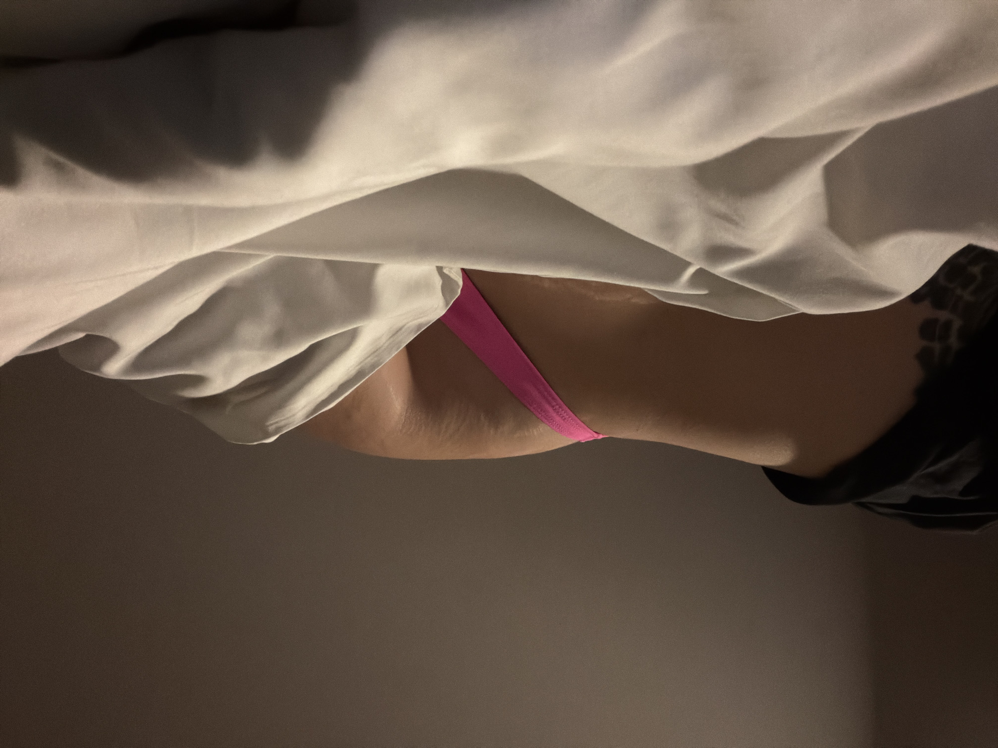 A close-up, low-angle shot shows a person lying in bed under a white sheet. A pink thong is visible, partially obscured by the sheet. The person is wearing a dark-colored shirt. The background is a plain, neutral-toned wall. The location and city are not discernible from the image.