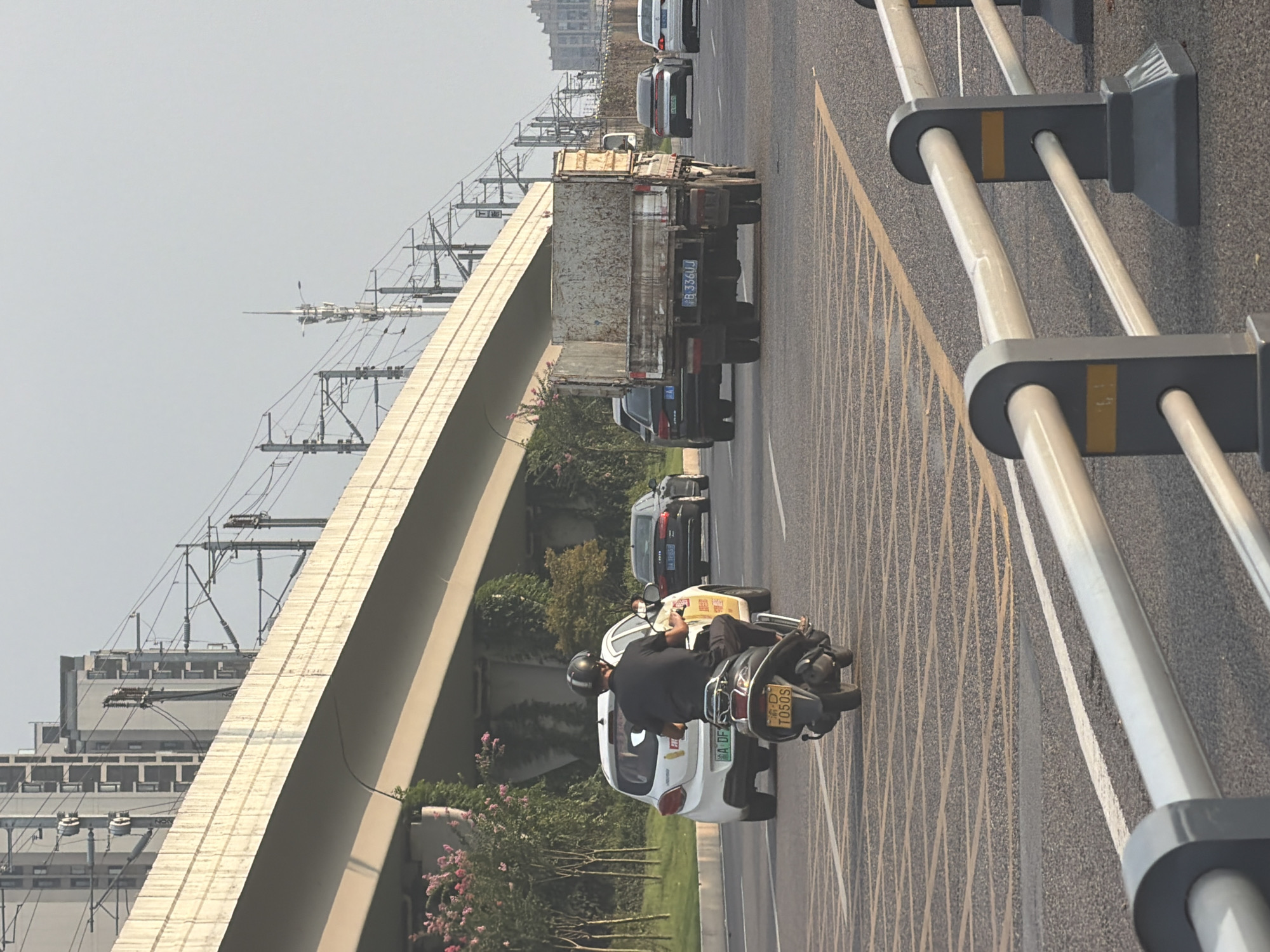 This image depicts a street scene in Jiangjin, China, under an elevated highway or bridge carrying what appears to be a railway line. The sky is a pale, hazy blue, suggesting it is daytime, possibly late afternoon due to the lighting.
In the foreground, a silver motorcycle with a dark-colored rider wearing a helmet is moving away from the camera. The motorcycle has a yellow license plate with black characters that read "T0505". To the left of the motorcycle, a white car is also moving away. This car has a license plate with green characters and a yellow character on the left. Behind the white car, a black sedan is visible.
Further down the road, a weathered, light-colored truck with a flatbed is moving away from the camera. Its license plate appears to be blue with white characters, reading "B 336UJ". To the right of the truck, a dark sedan is in the adjacent lane, also moving away. Beyond that, another white sedan is visible.
The road is paved with asphalt and marked with yellow and white lines. There are yellow grid markings on the road in front of the motorcycle. Along the left side of the road, there is a sidewalk with green vegetation, including some flowering trees with pink blossoms, and ivy growing on the concrete supports of the elevated structure. Metal railings, painted grey with yellow accents, line the right side of the road in the foreground.
In the background, tall, modern buildings with multiple windows are visible on the left, behind the elevated structure. Numerous electrical wires and poles associated with the railway line are prominent above the elevated structure. On the far right, more buildings are visible in the distance. The overall impression is one of urban infrastructure with active traffic flow.