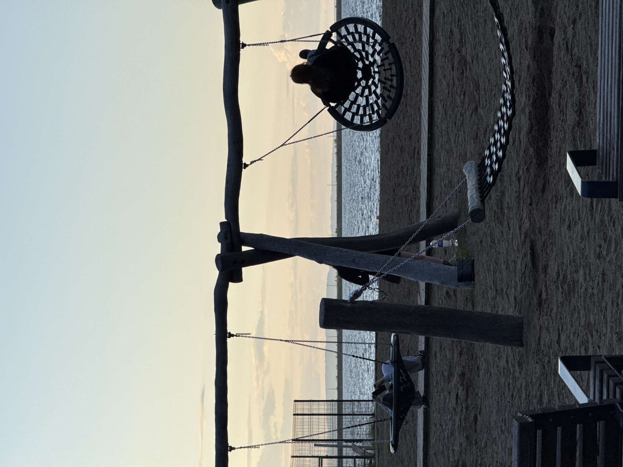 The image depicts a tranquil scene at a waterfront playground, likely during late afternoon or early evening. The sky, transitioning from a soft light blue at the top to a warm, pale yellow and orange near the horizon, backlights the entire scene, causing the foreground elements to appear in silhouette.
The primary subjects are two individuals enjoying swings. On the left, a person with visible hair is seated in a large, round "nest" swing, suspended by chains from a rustic wooden A-frame structure. To the right, another person is visible on a smaller, more traditional swing, also attached to the same or a connected wooden frame. Both figures are facing towards the distant body of water.
The setting is a sandy or dirt ground area that serves as the base for the swing set. Beyond the playground, a vast expanse of calm water, possibly a lake or large river like the Votkinsk Reservoir near Chaikovsky, stretches into the distance. A faint, distant shoreline with subtle land features is visible on the horizon line. In the mid-ground, near the smaller swing, a metal fence or railing is present. On the far right of the frame, parts of what appear to be wooden benches or seating areas are visible.
The overall mood is peaceful and serene, with the warm, fading light suggesting a calm end to the day. There is no visible text in the image.