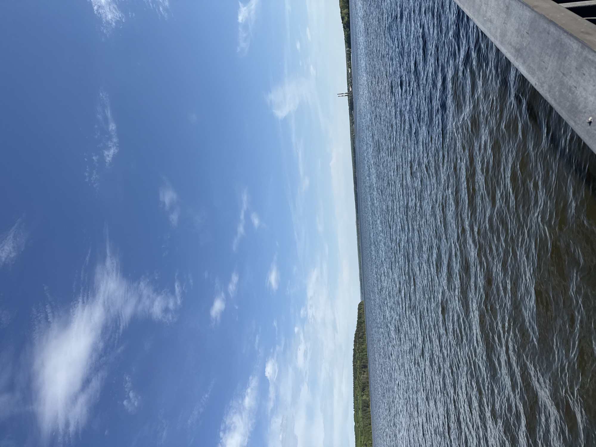 The image depicts a body of water, likely a river or lake, adjacent to a blue sky with scattered clouds. A strip of land with trees is visible on the far side of the water, and some industrial structures (possibly smoke stacks) are also discernible. The view is taken from a slightly elevated position, possibly a pier or boardwalk, with a portion of the structure visible in the upper right corner. While a specific location confirmation in the city of Chaikovsky, Russia is not possible without more context, the scene appears to be a common landscape associated with waterfront areas.