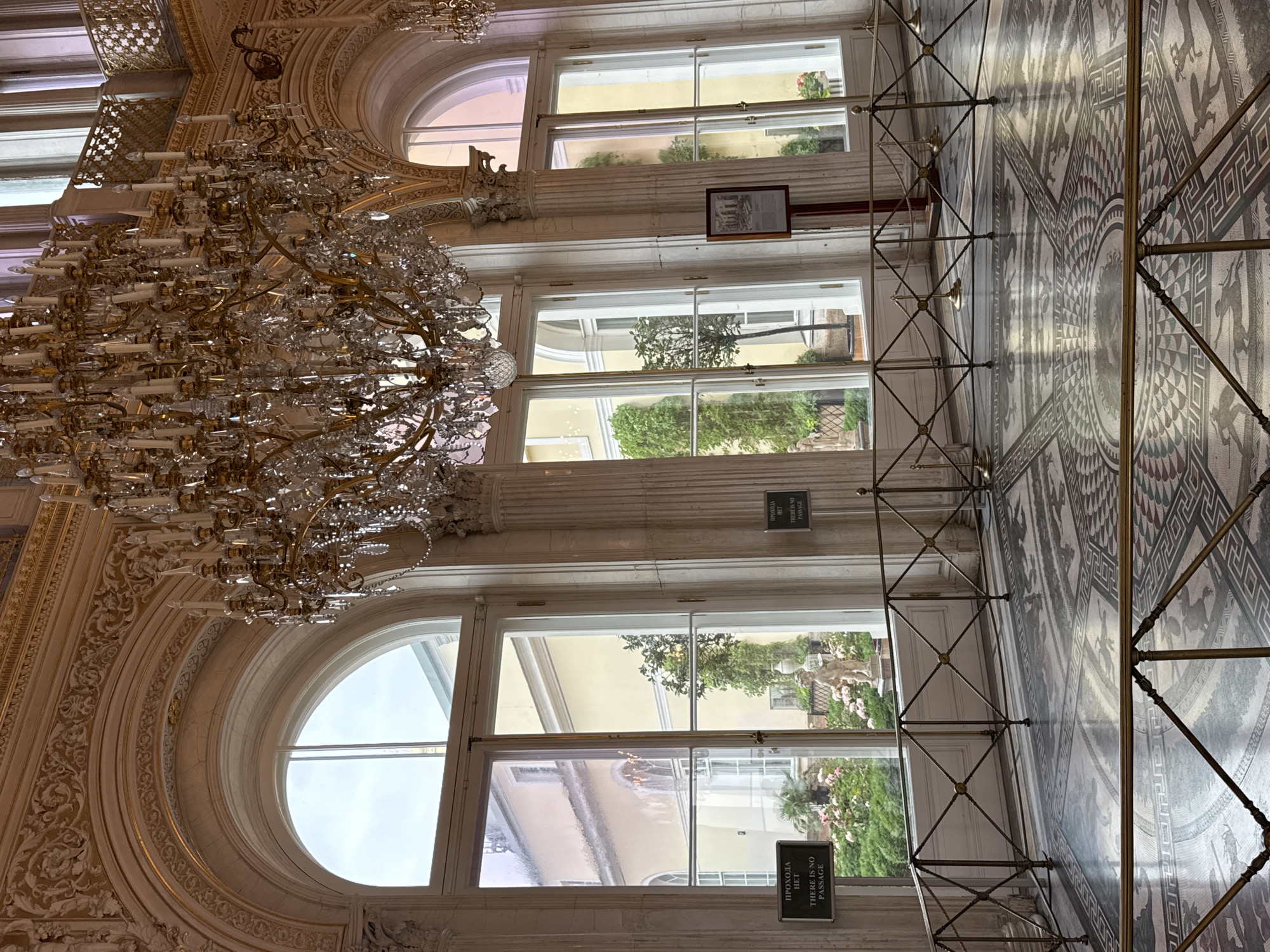 The image captures an opulent interior with a grand chandelier in the foreground. The chandelier is large and elaborate, adorned with numerous crystals and emanating a warm glow. To the right, a series of tall windows look out onto a courtyard or garden. The windows are divided into horizontal panes, and through them, one can see lush greenery, potted plants, and architectural elements, suggesting an outdoor space. The sunlight streaming through the windows casts dappled shadows on the ornate mosaic floor.
The mosaic floor is a prominent feature, displaying intricate patterns and figures in a grayscale palette. A metal railing system runs along the right side of the image, separating the interior space from the windows and floor.
The walls of the interior are decorated with elaborate stucco work and moldings, indicative of a historical and luxurious building. Above the windows, arched elements are visible, contributing to the grand architectural style.
Two small signs are visible on the walls near the windows, each with text in both Cyrillic and English. The English text reads "THERE IS NO PASSAGE."
The overall impression is one of grandeur and historical significance, likely a museum, palace, or other notable public building. The lighting suggests it could be daytime, with natural light augmenting the interior illumination. The scene is static, focusing on the architectural and decorative elements of the space.