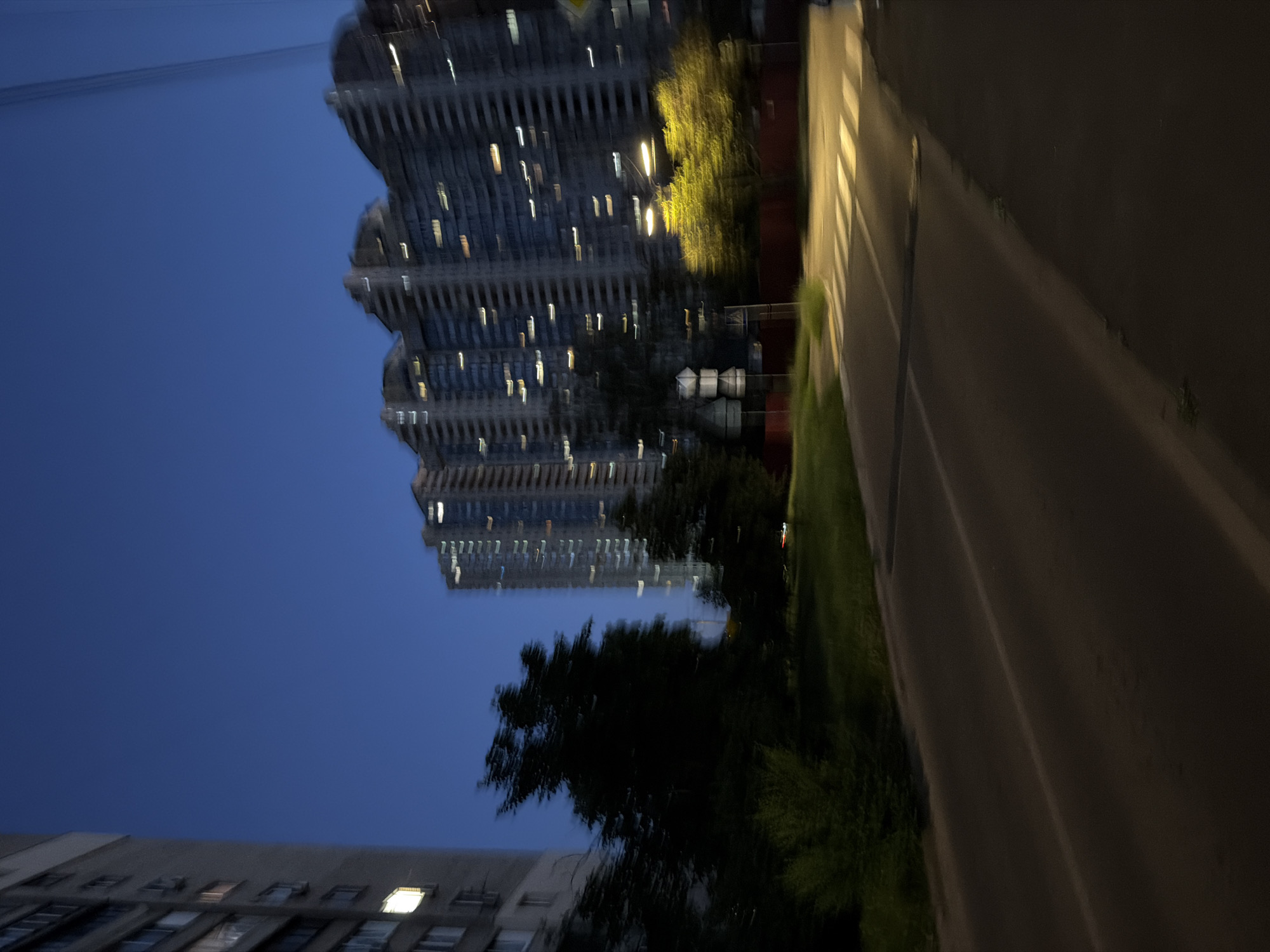 The image depicts a blurred urban scene at dusk or night in Kyiv, Ukraine. The sky is a deep blue. On the left, a tall, light-colored residential building with lit windows is partially visible. To the right of this building, dark trees and bushes are prominent. Further to the right, a large, multi-story residential building with many illuminated windows is the dominant feature, its silhouette softened by motion blur. In the foreground, a road runs diagonally across the image, with a pedestrian crossing marked by white stripes illuminated by streetlights. The overall impression is one of movement and low light conditions, characteristic of a late evening in a city.