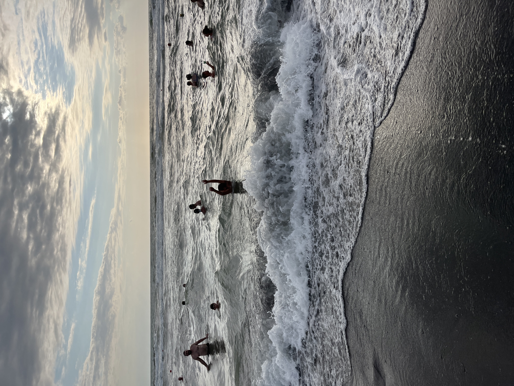 The image depicts a crowded scene at Ureki beach in Georgia, with numerous people bathing in the Black Sea. The water is choppy, with waves breaking on the dark, sandy shore. The sky is overcast with clouds, suggesting it may be late afternoon or early evening. Several individuals are visible in the water, some with their arms raised, possibly enjoying the waves or posing for a photo. The presence of many people indicates a popular recreational area.