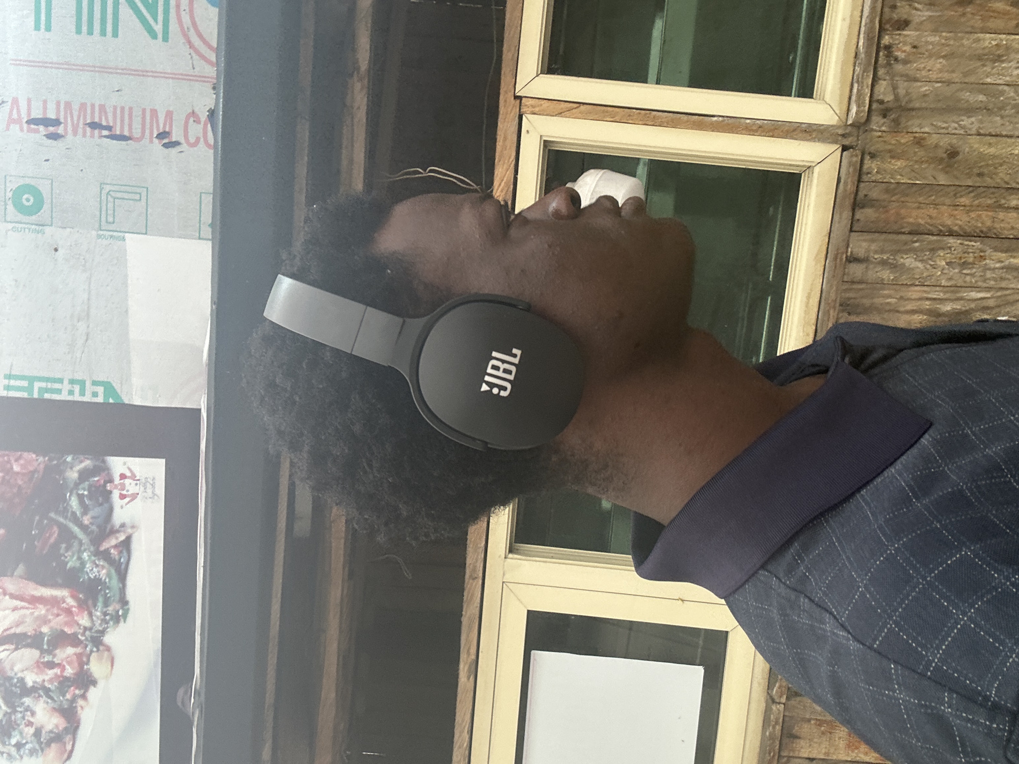 The image shows a young Black man wearing black JBL headphones and a dark blue collared shirt with a subtle plaid pattern. He is positioned in profile, facing to the right. Behind him, there are wooden planks forming a wall and windows with light-colored frames. In the upper left corner of the image, a framed picture of food is visible. The overall scene appears to be outdoors, possibly in a casual setting within the city of Badagry, Nigeria.