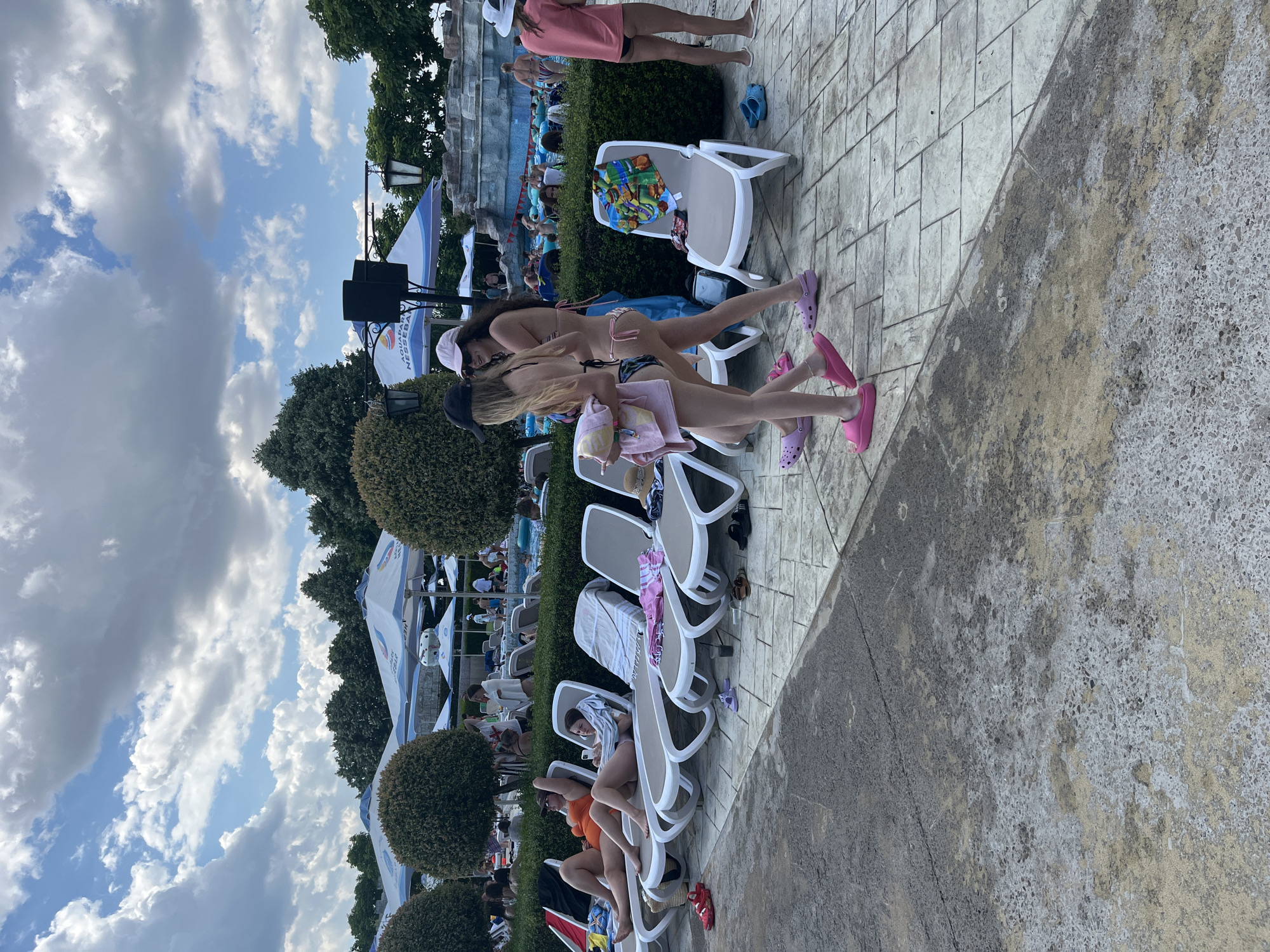 This is a medium shot of a poolside area at a water park, possibly located in Ravda, Bulgaria, given the "Aquapark Nessebar" branding visible on a banner. The scene depicts a sunny day with scattered clouds in a blue sky. In the foreground, two young women in bikinis are walking, one carrying towels. They are wearing casual footwear, pink slides and purple slides respectively. To their left, several lounge chairs are occupied by people sunbathing or relaxing, some under large umbrellas. One woman in an orange swimsuit is lying on a lounge chair, looking at her phone. Further back, a water slide structure is visible, with people enjoying the water and a lazy river. The ground is paved with stone tiles and concrete. Several towels are draped on lounge chairs and on the ground, suggesting a busy atmosphere. The overall environment appears to be a recreational facility designed for relaxation and aquatic activities.