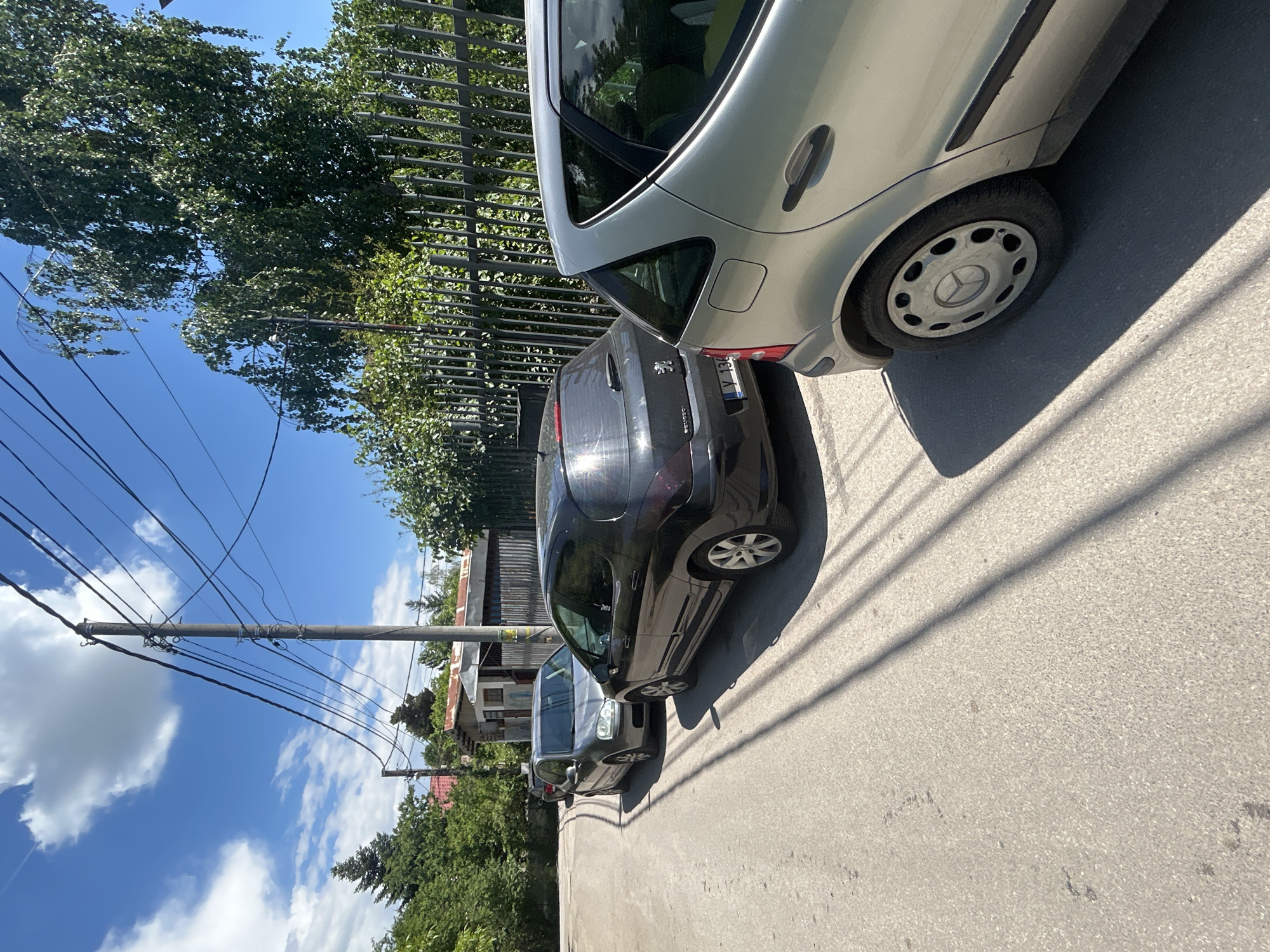 The image depicts a street scene in Sofia, Bulgaria. Two cars, a silver hatchback and a dark grey/black hatchback (likely a Peugeot 307 based on its rear design), are parked closely together on the right side of the road. The silver car is positioned at the front, slightly angled inwards. A third car, dark in color, is parked further down the road. A metal fence runs alongside the street, partially obscured by trees. A utility pole with power lines extends across the sky. The sky is blue with scattered white clouds. The road appears to be asphalt, with shadows cast by the cars and overhead wires. The scene is captured in daylight with clear visibility. Typical Bulgarian houses are seen in the background.