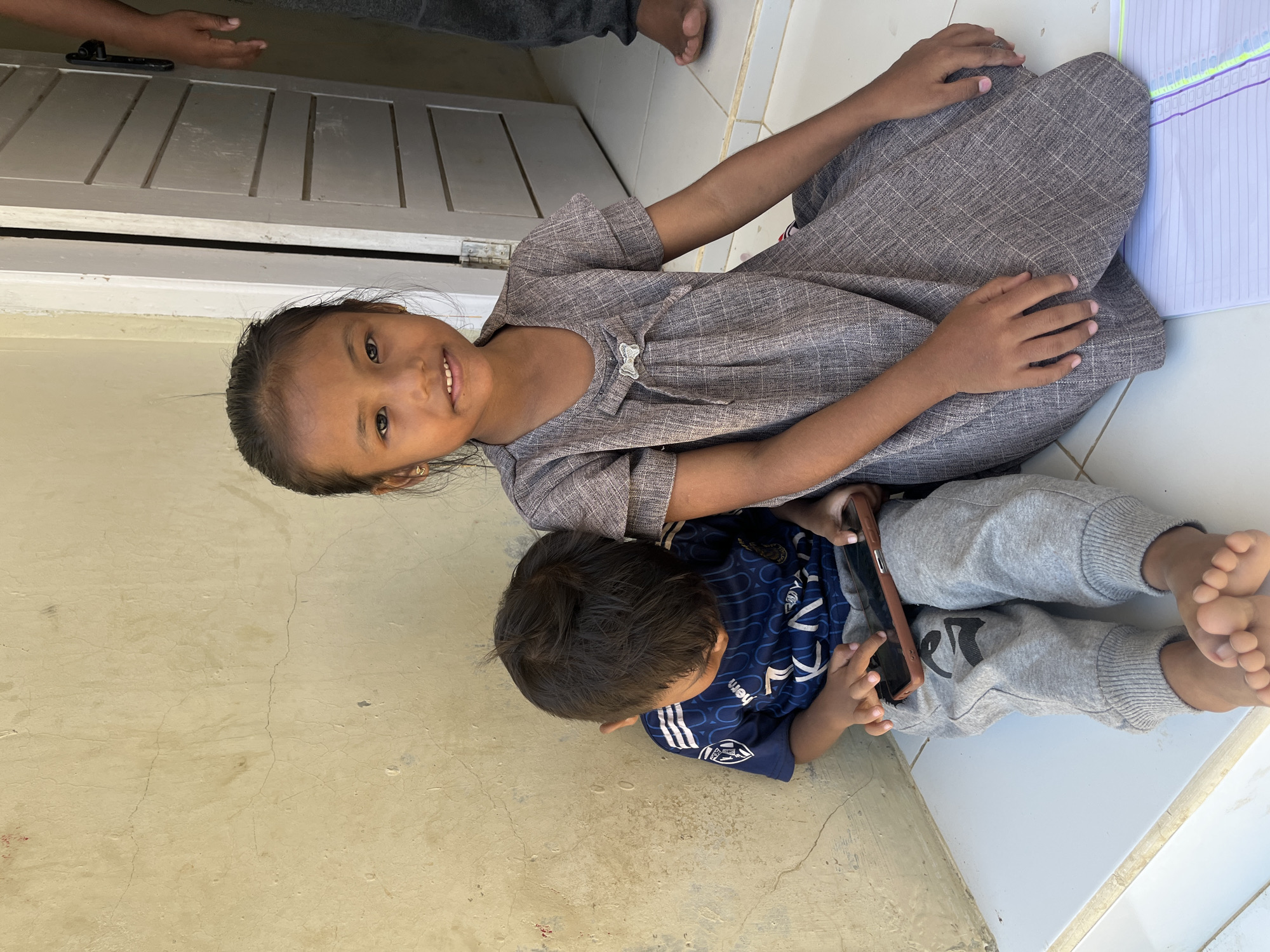 In Geunteng, Indonesia, two children are seated outdoors on a tiled surface next to a pale yellow, textured wall. A young girl with dark hair pulled back is on the right, looking towards the camera with a slight smile. She wears a grey, checkered dress with a decorative bow at the neckline. To her left, a younger boy is looking down at a smartphone he holds in his hands. He wears a dark blue t-shirt with white stripes and lettering, and light grey sweatpants. His feet are visible, and he is barefoot. Behind the children, a light-colored wooden door is partially ajar, revealing a dark interior. A third person's legs and feet are visible to the right of the door, wearing dark trousers. The environment suggests a casual, outdoor setting, possibly near a dwelling.