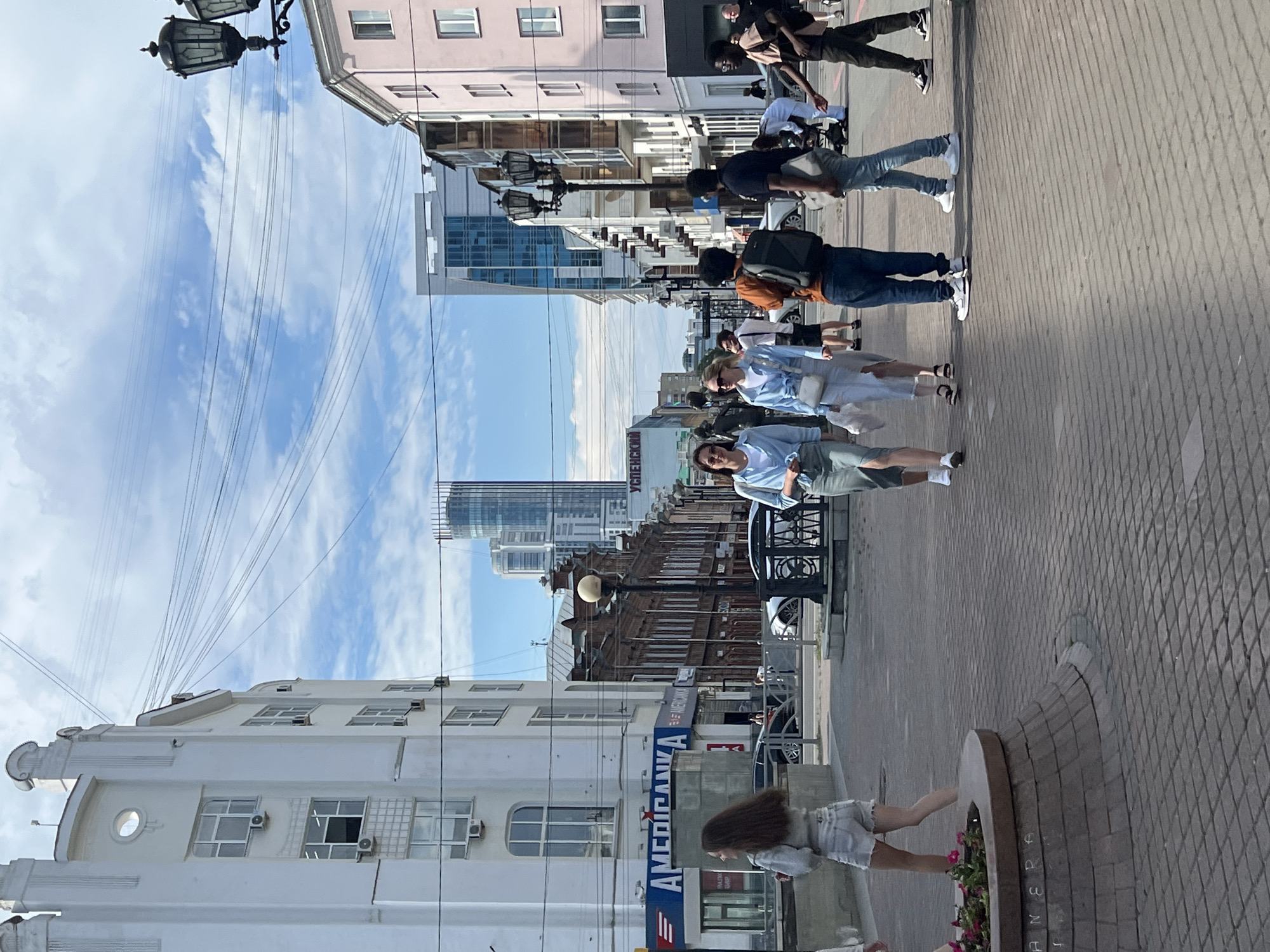 The image depicts a street scene in Yekaterinburg, Russia, specifically on Vaynera Street. A group of pedestrians is walking along a paved sidewalk. In the foreground, to the left, a woman in denim shorts and a light-colored top is walking away from the camera. To her right, two women are walking towards the camera, one in a light blue top and a light-colored skirt, and the other in a similar blue top and a white skirt. Further ahead, several other individuals are walking in various directions.
The environment includes buildings lining the street, with architectural styles ranging from older, ornate brick structures to modern, glass-fronted buildings. One prominent building on the left features the signage "AMERICANKA". Overhead, a complex network of wires crisscrosses the sky, characteristic of urban infrastructure. Streetlights and decorative lampposts are also visible. A round planter with flowers is situated on the left side of the sidewalk. The sky is partly cloudy, indicating daylight. The overall impression is a bustling urban environment on a clear day.