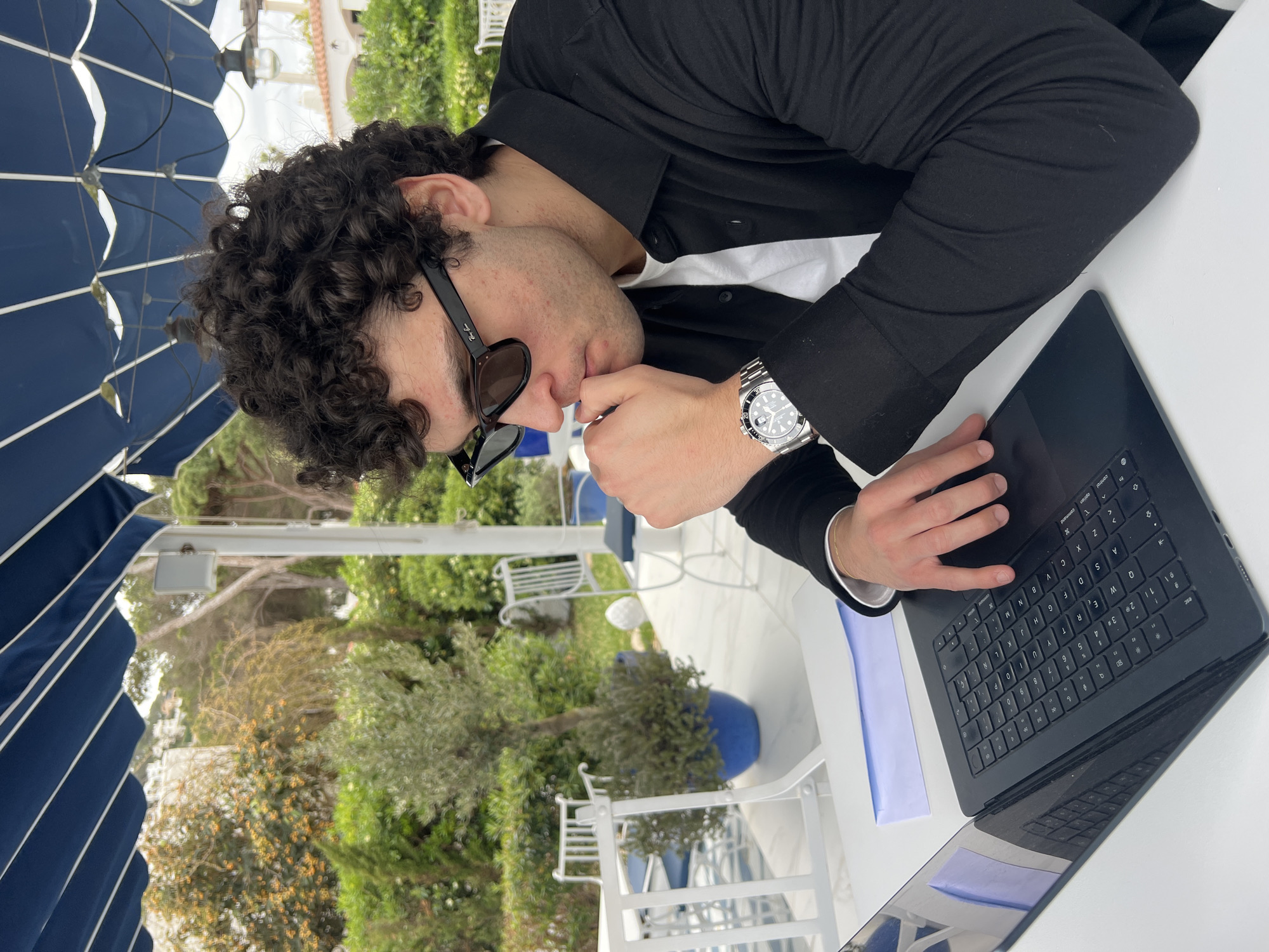 A person with dark curly hair is sitting at a white table under a blue and white striped umbrella. The person is wearing sunglasses, a black shirt, and a white t-shirt underneath. They are also wearing a silver watch. The person is using a black laptop. A blue folder is next to the laptop. In the background are green trees and plants, as well as white chairs and blue plant pots. The scene is likely in Santa Eulària des Riu, Spain.
