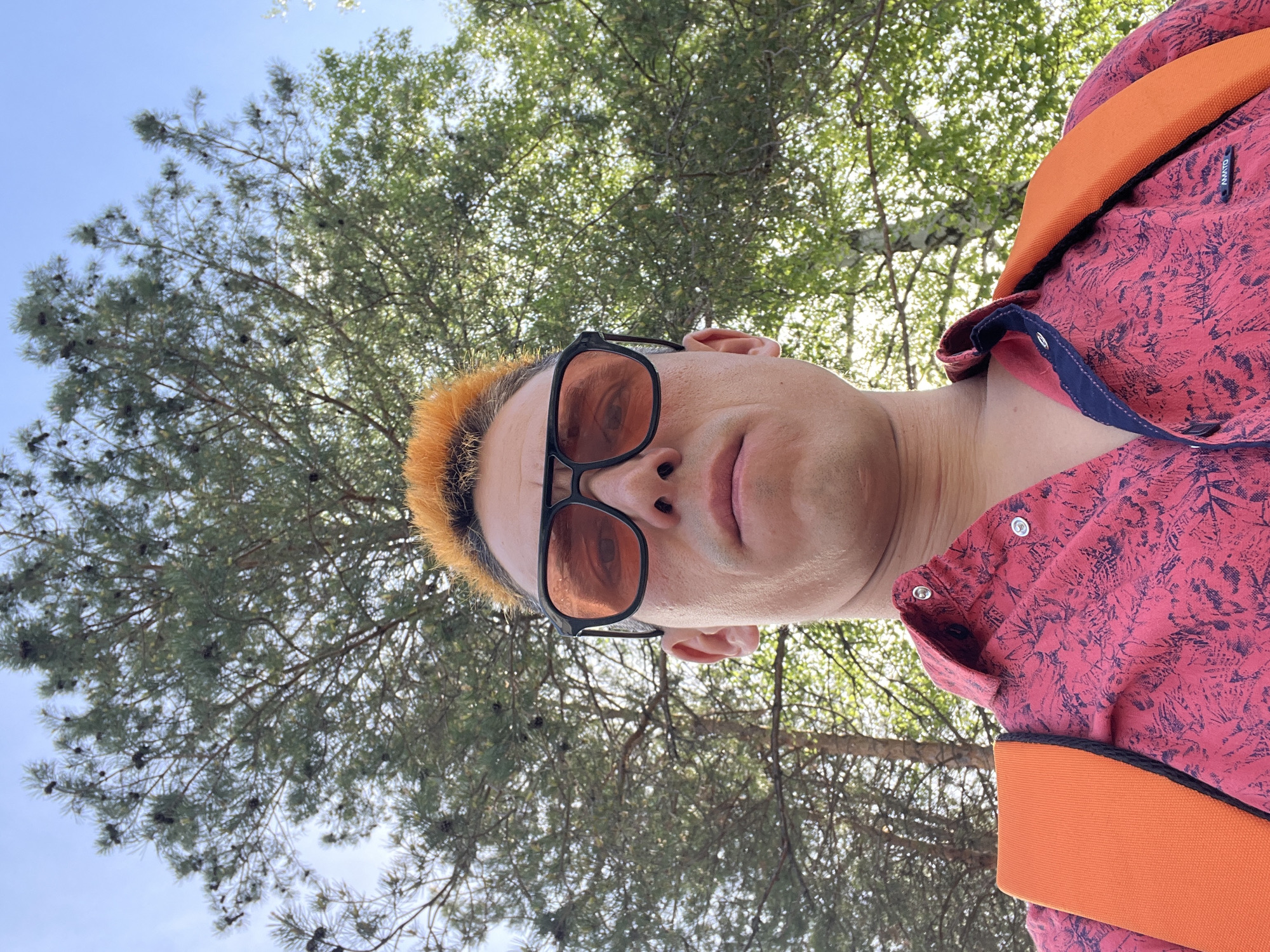 A low-angle, outdoor portrait shows a person against a backdrop of trees and sky. The person has pale skin, short, orange hair, and is wearing black-framed sunglasses with orange lenses. They are wearing a red patterned shirt with an orange strap running over the shoulder. Behind the person, trees with dense green foliage are visible, some branches reaching into the sky. The sky is a pale blue color. The location is in the city of Chaikovsky, Russia.