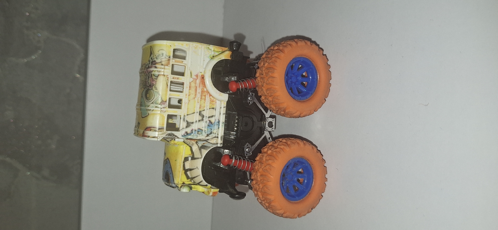 A toy monster truck, resembling a school bus with large orange tires and blue rims, sits on a white surface. The toy is yellow and white with graffiti-style graphics. Its suspension system, with red springs, is visible. The background is out of focus, showing a grey, textured surface with a light reflection at the top. There are no people or animals present in the image, and there is no information suggesting a location in Bila Tserkva, Ukraine.