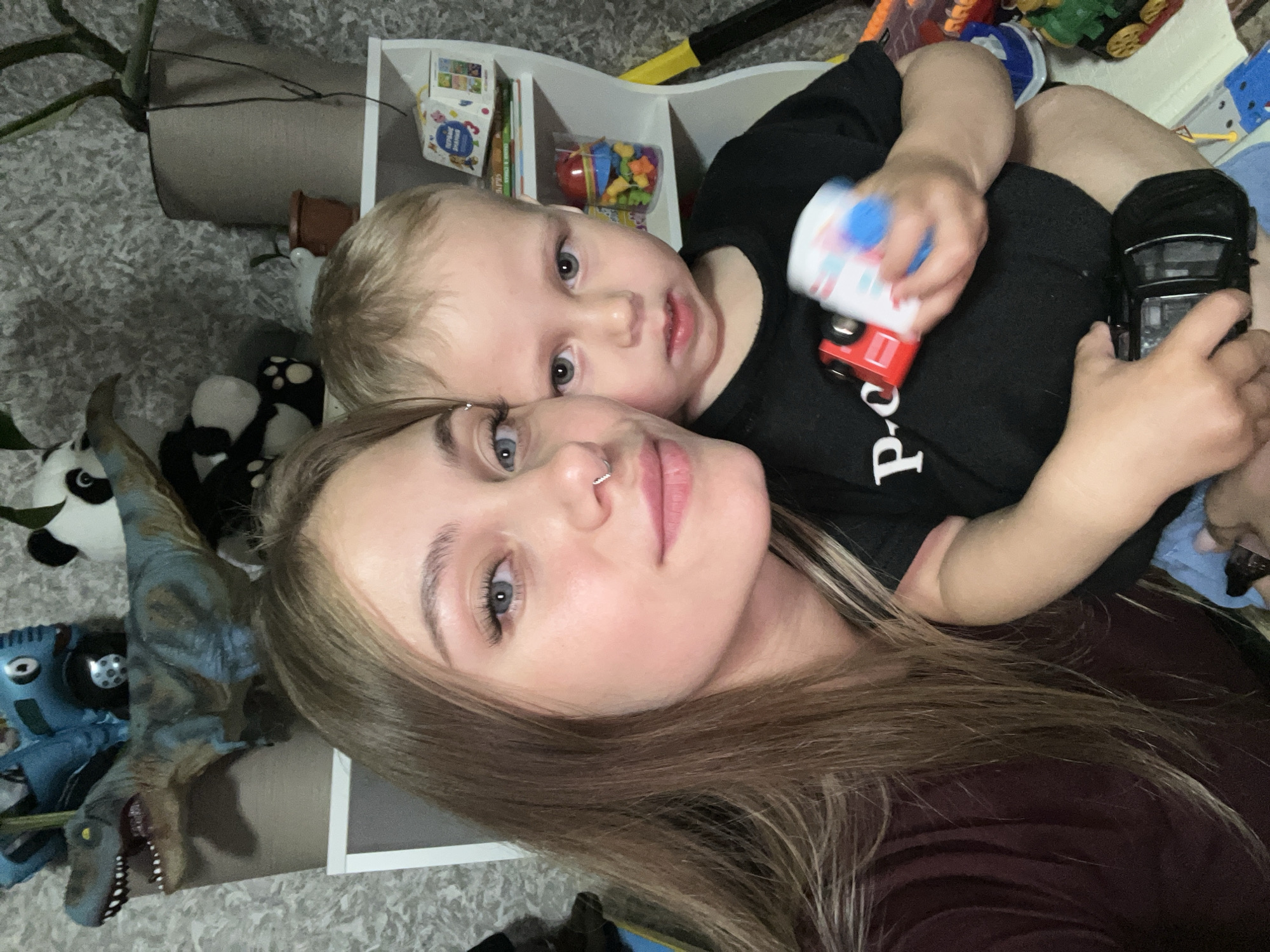 The scene depicts a young woman holding a small child within a residential interior identified as being in the city of Фоки, Russia. The woman, positioned on the left, has long, straight brown hair, blue eyes, and is wearing a silver septum piercing in her nose and a dark maroon-colored top. Her left arm is around the child. The child, held by the woman and positioned on the right, appears to be a toddler with short blonde hair and grey-blue eyes. The child is wearing a black t-shirt with a white partial "P-o" text visible on the left chest and blue shorts. The child is holding a small red toy truck in their left hand and a cylindrical white and blue toy in their right hand, with a black toy car held by their right thigh.
The background reveals a textured, light grey wall covering. A white shelving unit is present behind them, containing various items. On the top shelf, from left to right, are a realistic-looking blue-grey toy dinosaur, several deflated or partially inflated blue balloons, a black and white panda plush toy, and a large plant in a tall, light brown pot. Below the plant, on a lower shelf, is a smaller brown pot. On a middle shelf, a box labeled with Cyrillic text ("ПЕРВЫЕ ЗНАНИЯ" - First Knowledge) and a clear container filled with colorful building blocks or small toys are visible. To the right, a yellow and black object, possibly part of a toy, stands against the wall.