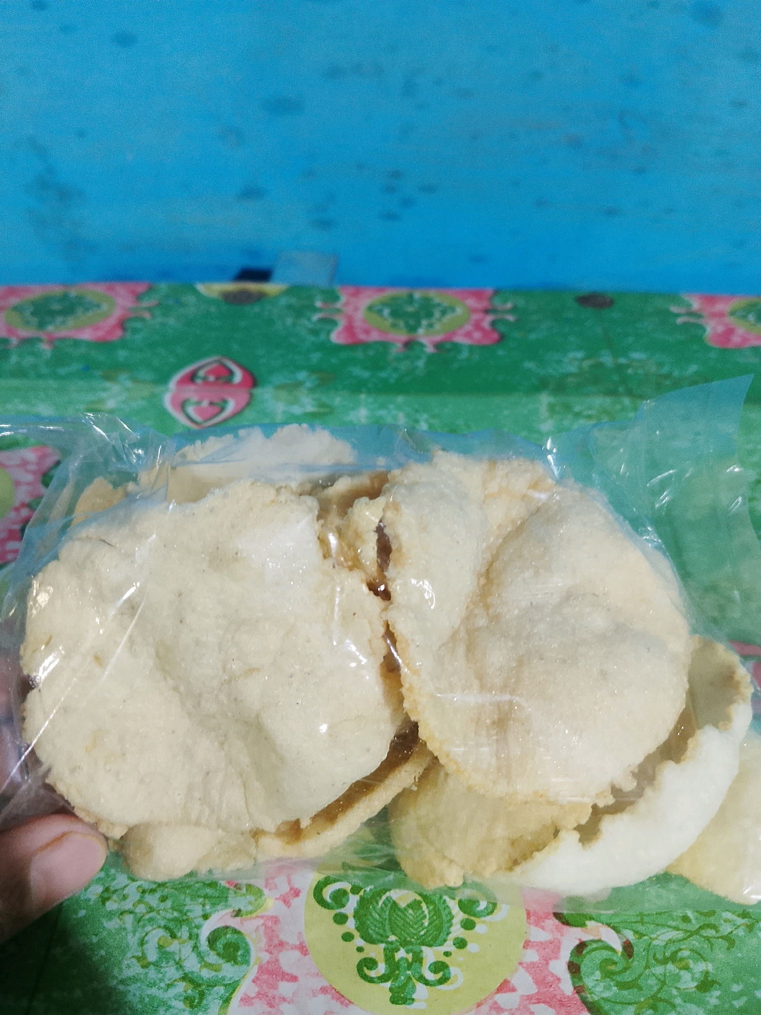 In Pelaihari, Indonesia, a close-up shot captures a bag of light-colored crackers, possibly krupuk, resting on a patterned surface with green, pink, and yellow designs. The crackers are partially obscured by clear plastic packaging, with some pieces showing signs of breakage. A person's thumb is visible on the left side of the frame, near the bottom of the bag. The background consists of a textured blue surface, possibly a wall, with some darker speckles.