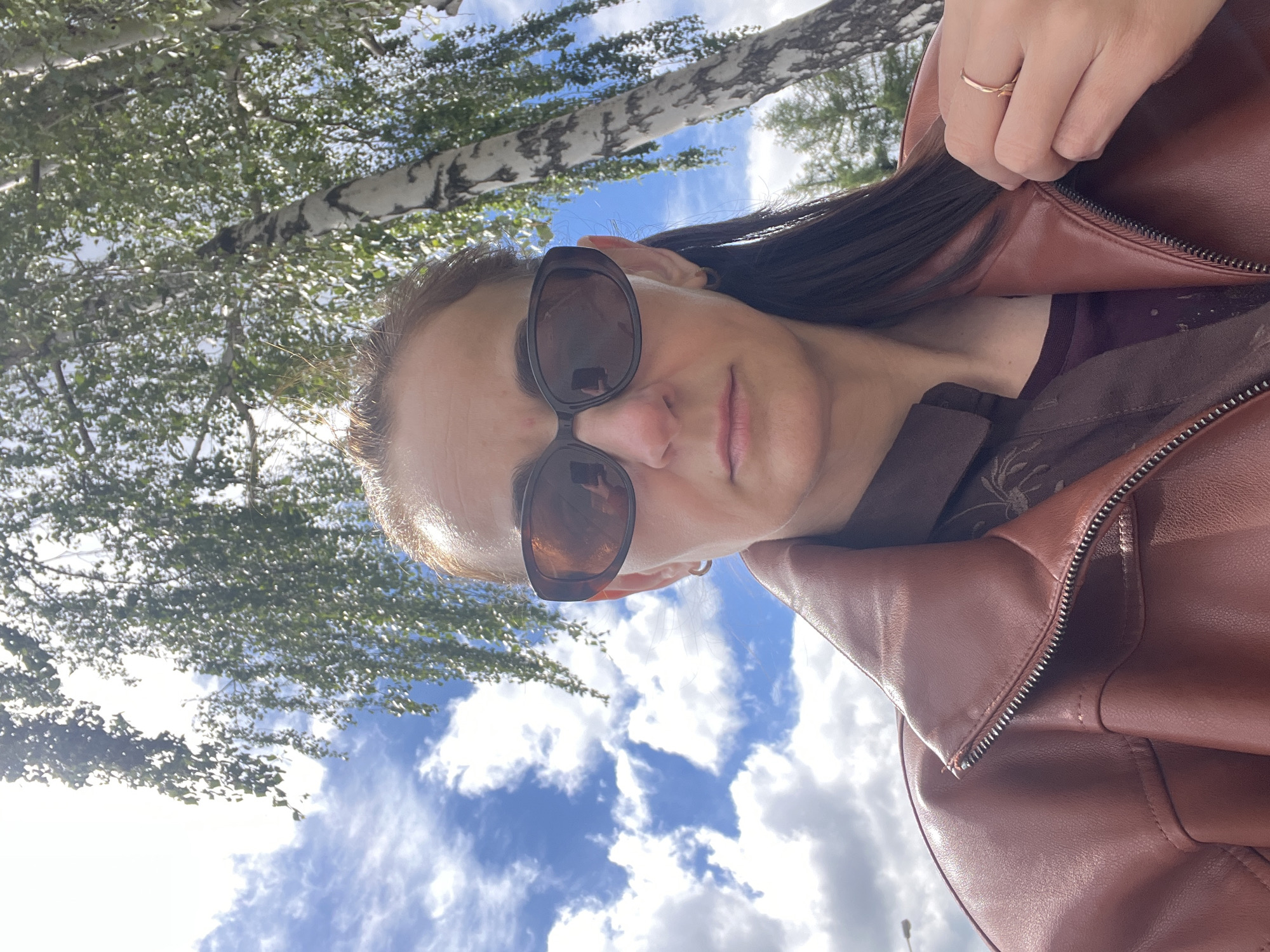 A selfie taken outdoors shows a woman wearing sunglasses and a brown leather jacket partially unzipped. In the background, there are trees with white bark and green leaves against a blue sky with white clouds. The location is unspecified within the city of Unknown, Unknown. The woman is holding the edge of her jacket, suggesting she may be in the process of opening or adjusting it.