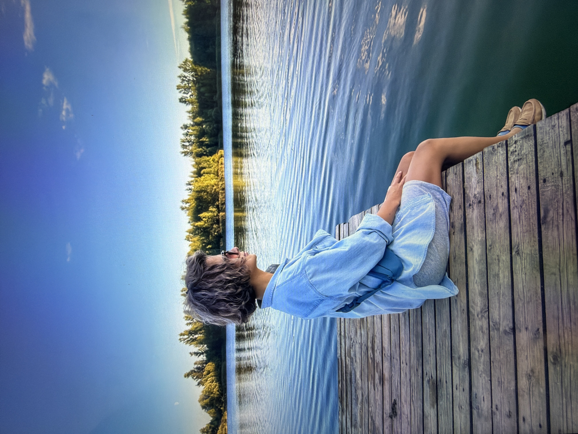 The image shows a woman sitting on a wooden pier, facing away from the camera and looking out at a calm lake. She has short, curly hair and is wearing a light blue denim jacket over a light-colored dress and grey shorts. A small blue bag is slung across her chest. Her legs are dangling over the edge of the pier, with her feet resting on the wooden planks. The lake is still, with ripples reflecting the blue sky and the green trees on the opposite shore. The sky is clear with a few wispy clouds, and the sun is casting a warm glow on the scene. This is an outdoor scene, likely in a natural setting, with no indication of the city of Kotelniki, Russia.
