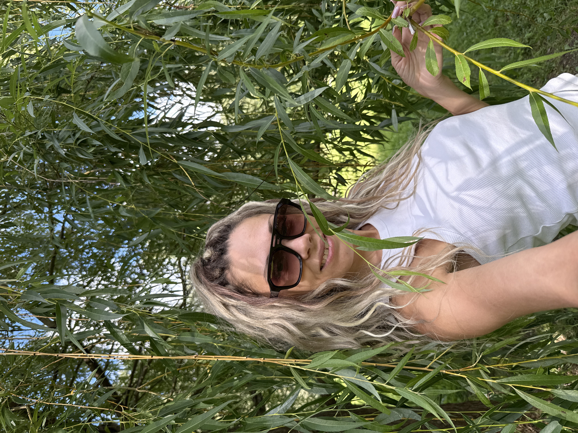 In Rusanovo, Russia, a woman with blonde, wavy hair and sunglasses smiles at the camera while standing amidst the drooping branches of a weeping willow tree. She is wearing a white ribbed tank top and her right hand is visible, reaching out to touch a branch. The background consists of lush green foliage and a glimpse of a blue sky.